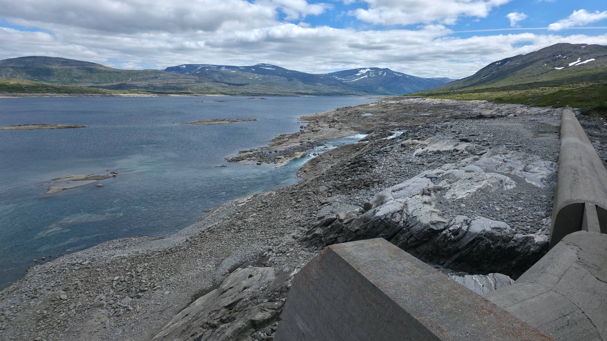 Betongdemning ved et vann med lav vannstand og fjell i bakgrunnen under en blå himmel. (Bildebeskrivelsen er laget av en KI-tjeneste)