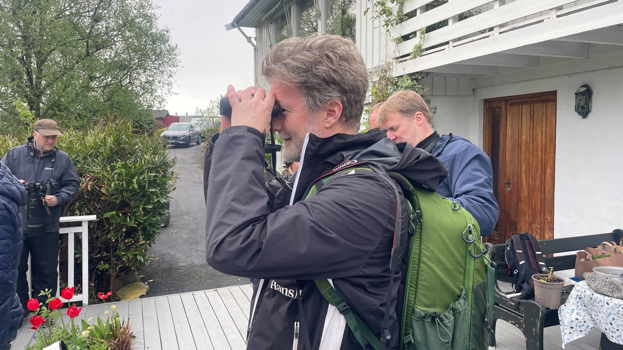 Menn står på ein terrasse og kikar ut i skogen med kikkertar og fotokamera med lange linser