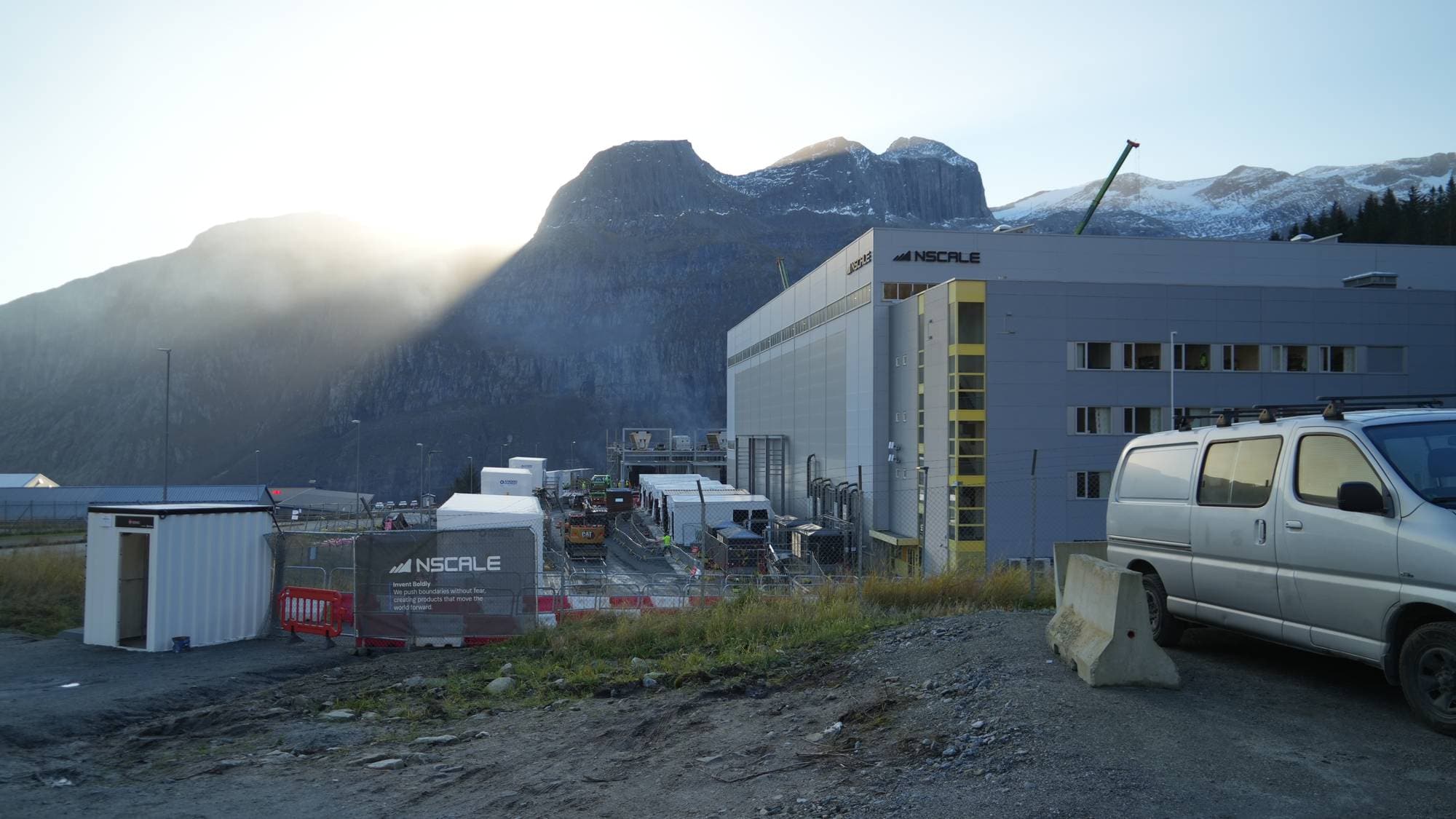EN bil parkert foran et stort industribygg, med fjell i bakgrunnen. 