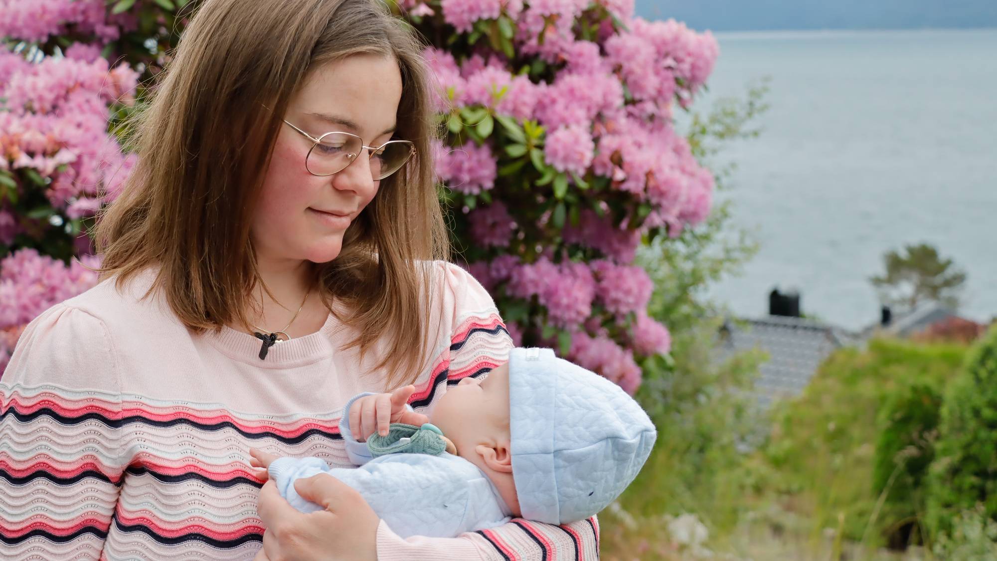 En kvinne med briller holder en baby i blå klær. Bak dem er det blomstrende rhododendronbusker. I bakgrunnen skimtes et hav og hus. (Bildebeskrivelsen er laget av en KI-tjeneste)