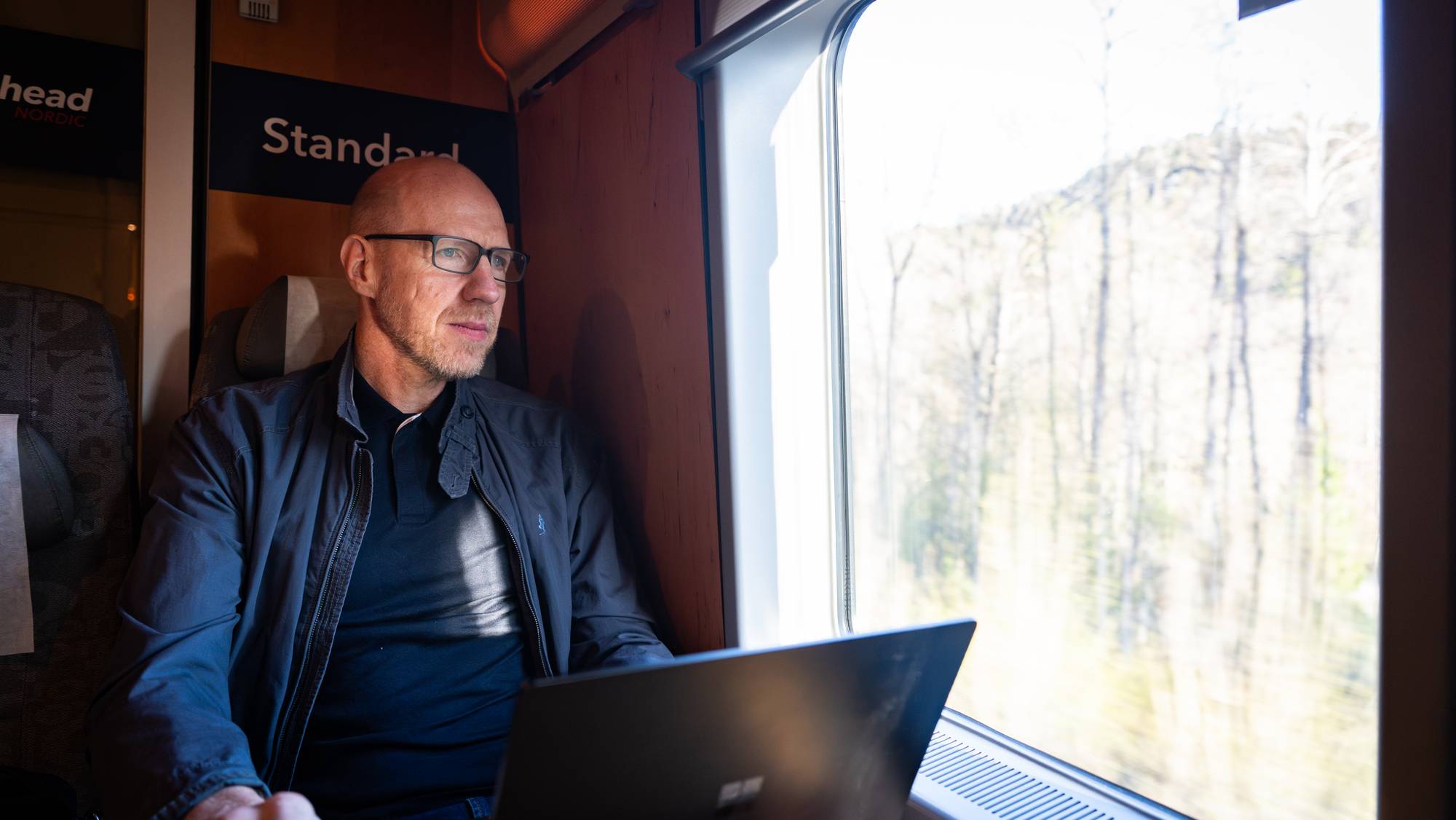 En mann sitter ved et togvindu og ser ut. Han har på seg briller og en jakke. En bærbar datamaskin står foran ham på et bord. Utenfor vinduet er det trær og vegetasjon. (Bildebeskrivelsen er laget av en KI-tjeneste)