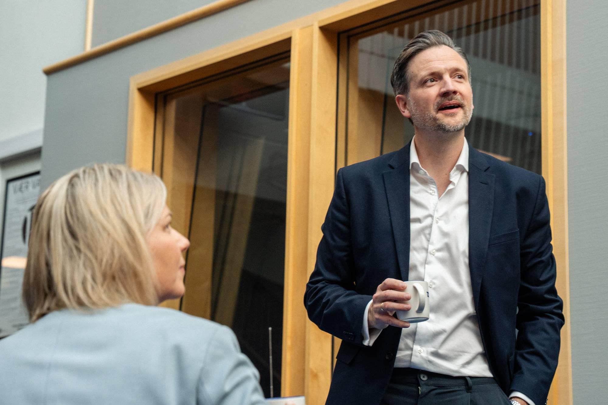 En mann i mørk dress og hvit skjorte holder en kaffekopp. En kvinne i lyseblå dress sitter med ryggen til. De befinner seg i et rom med lyddempende vegger og glassvegger. Bak dem er det montert lyddempende paneler. (Bildebeskrivelsen er laget av en KI-tjeneste)