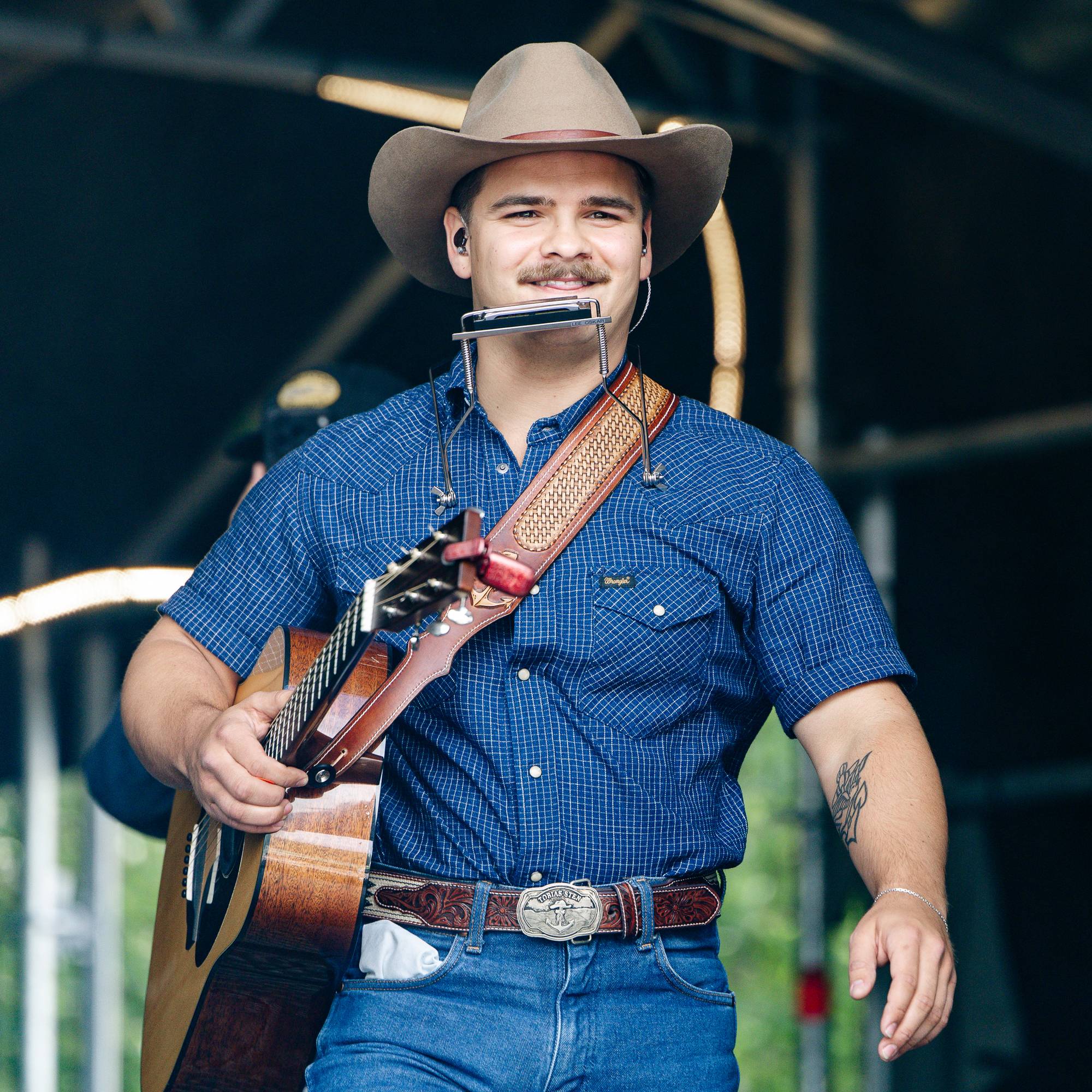 En mann med cowboyhatt spiller munnspill mens han holder en gitar. Han har på seg en blårutete skjorte og jeans. Mannen står på en scene under en utendørs konsert. Bak ham er det en mørk bakgrunn med lys og sceneutstyr. (Bildebeskrivelsen er laget av en KI-tjeneste)