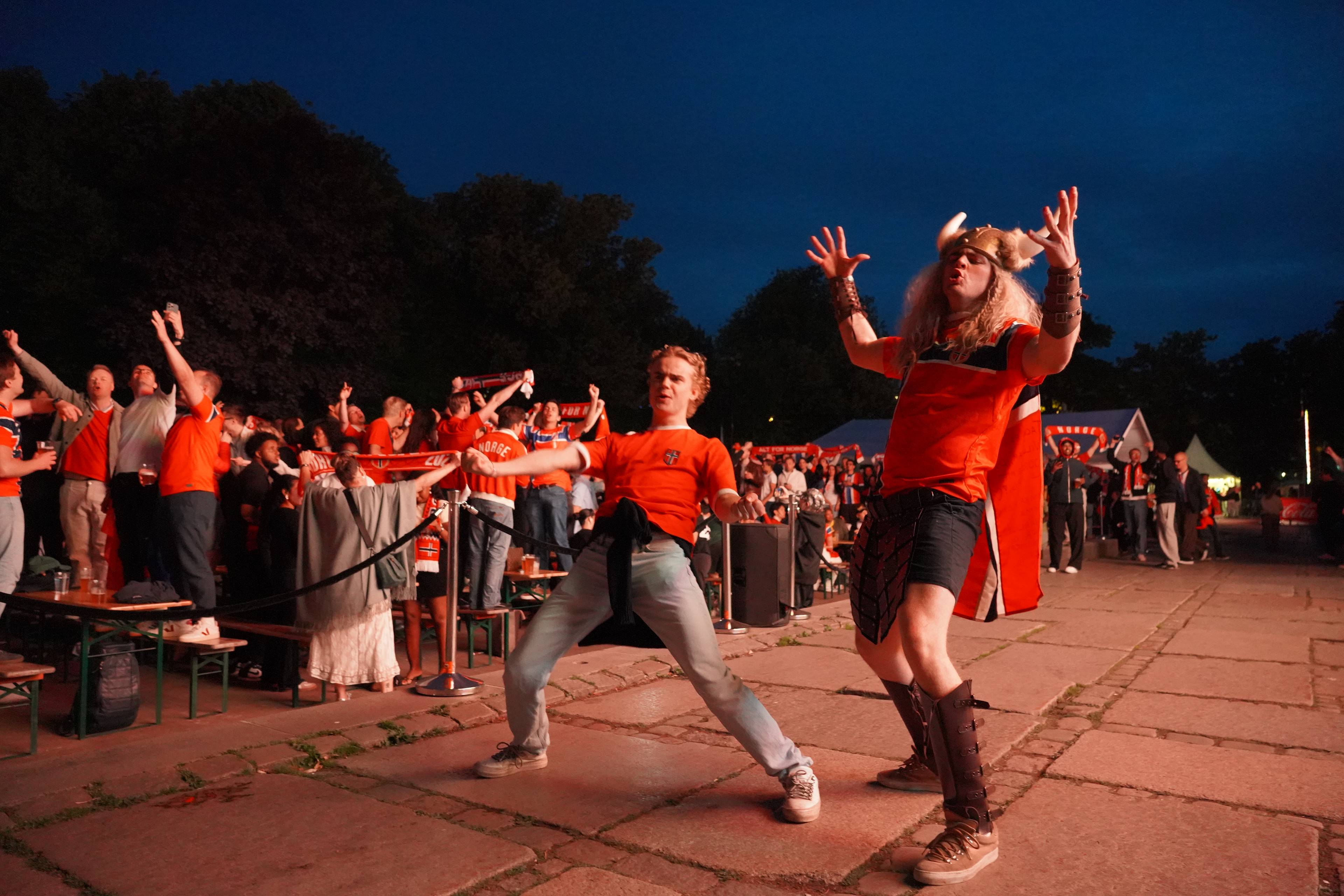 Publikum ser ut til å feire og danse foran en scene utendørs. To menn er i forgrunnen, den ene kledd i det som ser ut som en vikingdrakt. Det er flere mennesker bak dem, mange kledd i oransje og holder opp skjerf.