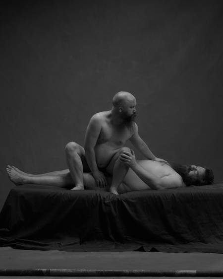 To nakne menn med skjegg på en mørk seng. Den ene mannen ligger utstrakt på senga med hånda på den andres kne. Den andre sitter sidelengs på huk med rumpe mot skritt på den andre mannen. Den ene hånda hans er mellom beina sine, mens den andre er på partneres bryst. De ser på hverandre.