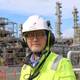 Equinor Bernt Tysseland, Equinors anleggsdirektør på Mongstad, står med hjelm og gul jakke. I bakgrunnen to høge tårn og delar av Mongstadanlegget. Han har lyst skjegg og briller.