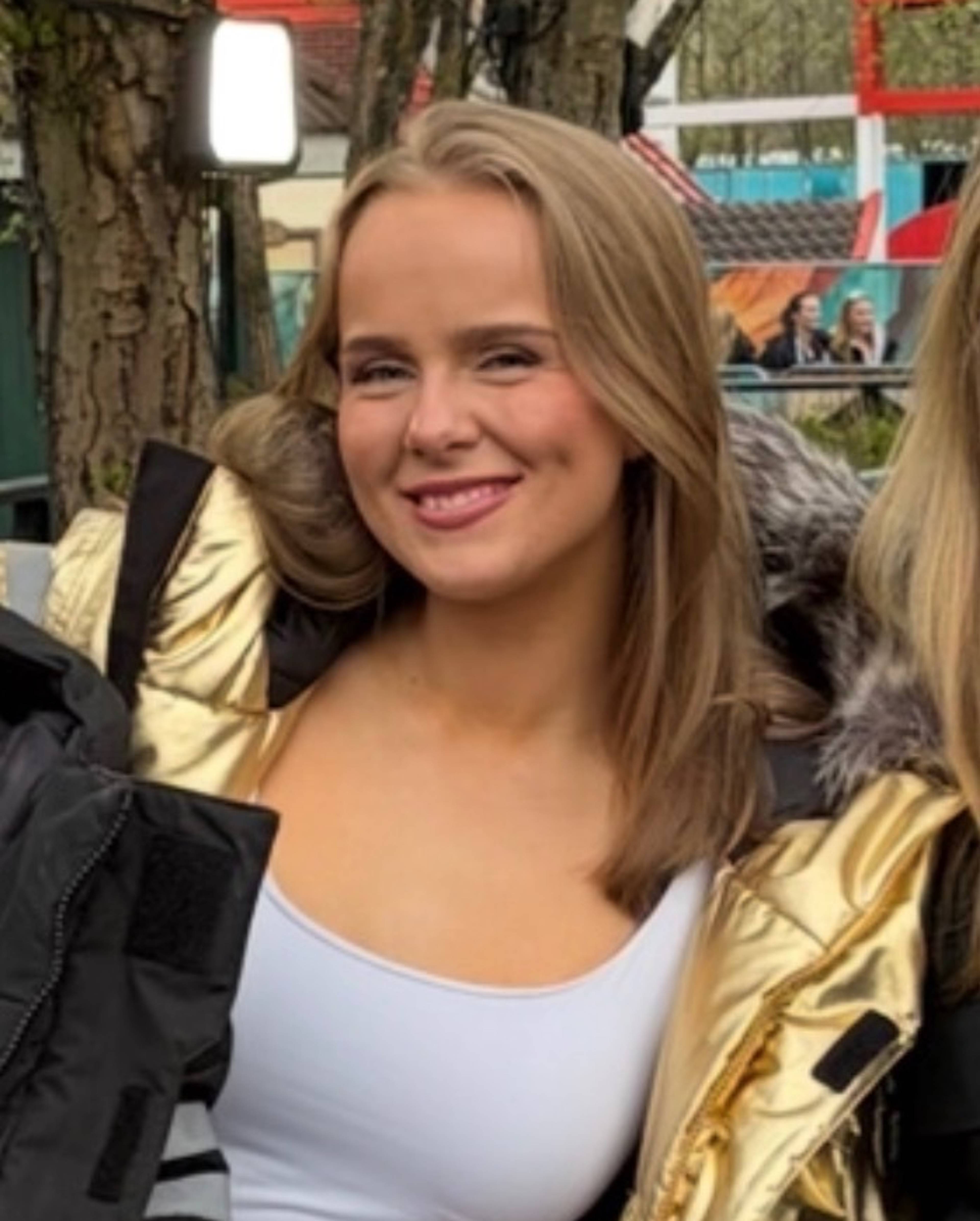 En jente i en gullfarget jakke smiler til kameraet. Hun har brunt hår og en hvit singlet under jakken.
