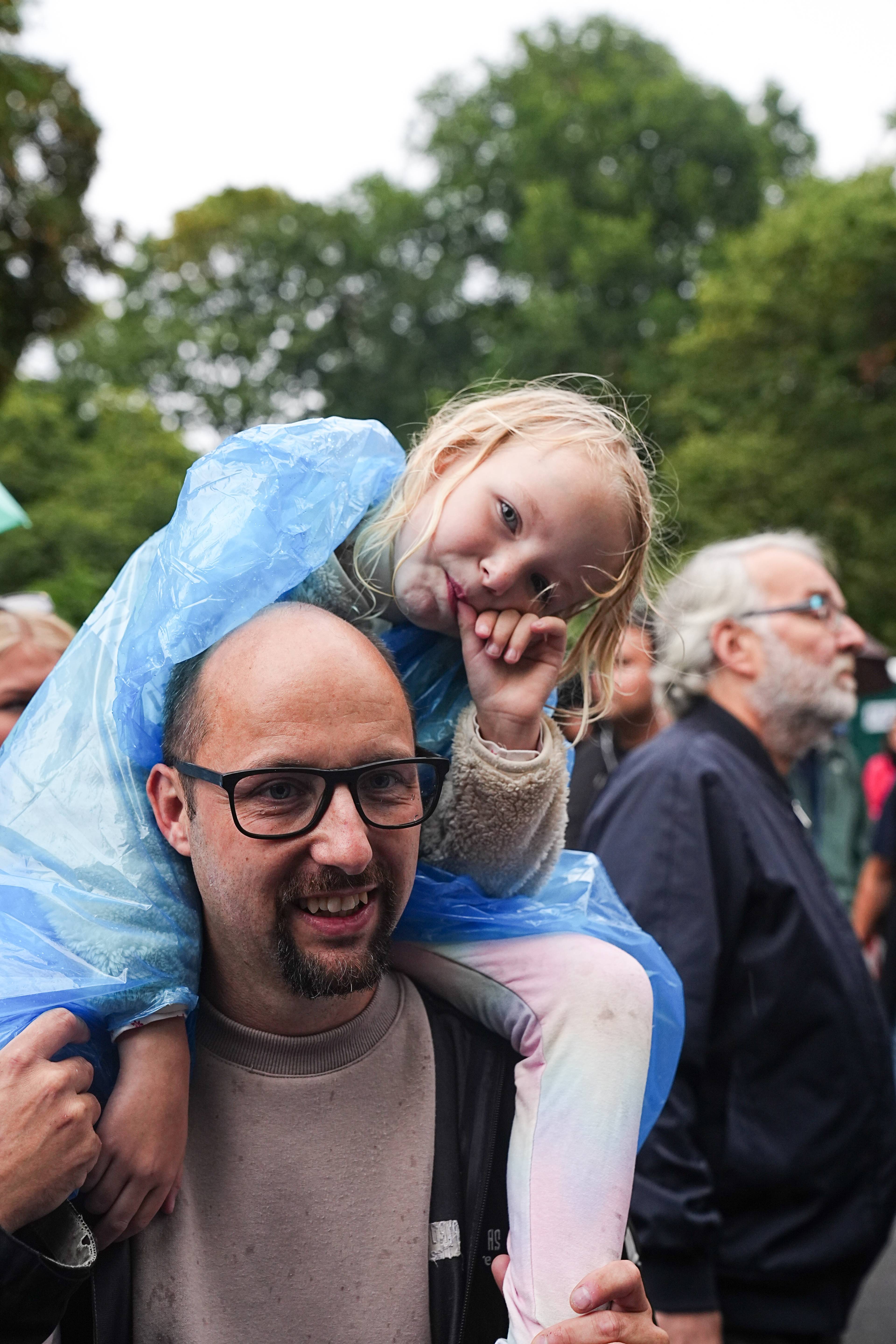 En mann bærer en liten jente på skuldrene under en blå plastregnponcho i et grønt uteområde. (Bildebeskrivelsen er laget av en KI-tjeneste)