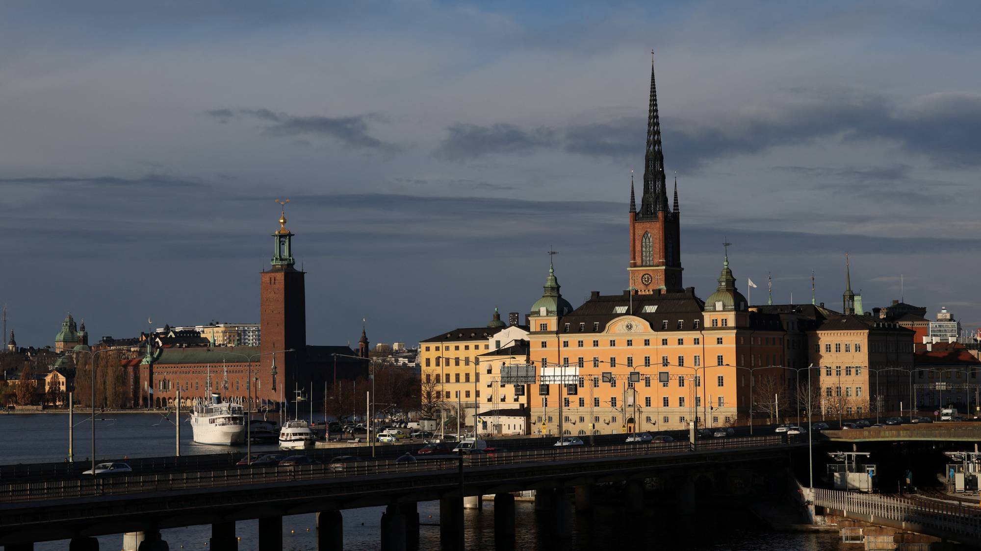 Bilde av Stockholms stadshus og Riddarholmskyrkan. En bro med biltrafikk går over vannet foran bygningene. Det ligger skip i havnen. (Bildebeskrivelsen er laget av en KI-tjeneste)