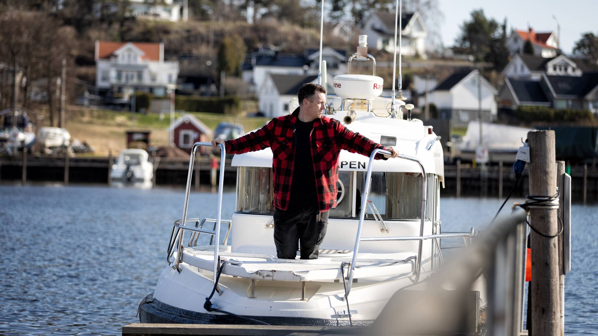 En mann står foran i båten og ser utover sjøen. Båten står inntil en brygge.