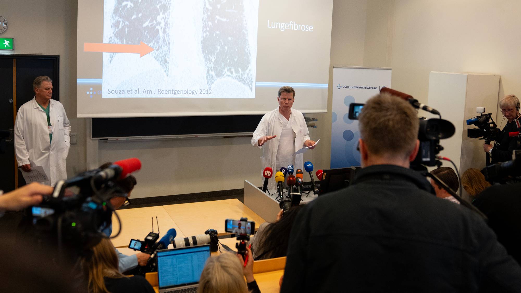 En lege holder en pressekonferanse foran en skjerm med røntgenbilder av lunger og teksten "Lungefibrose". En annen lege står til venstre. Flere mikrofoner og videokameraer er rettet mot legen ved podiet. En banner for Oslo Universitetssykehus er synlig i bakgrunnen. (Bildebeskrivelsen er laget av en KI-tjeneste)