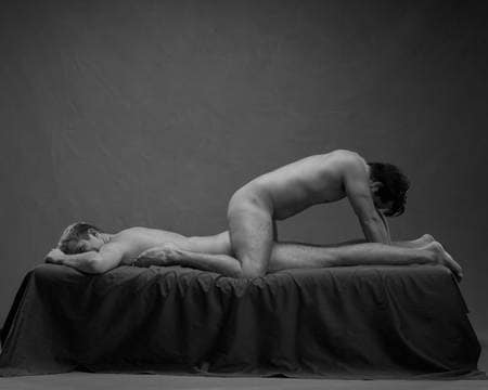En naken mann ligger på magen i senga. En annen naken mann står på kne over ham med hode mot føttene hans