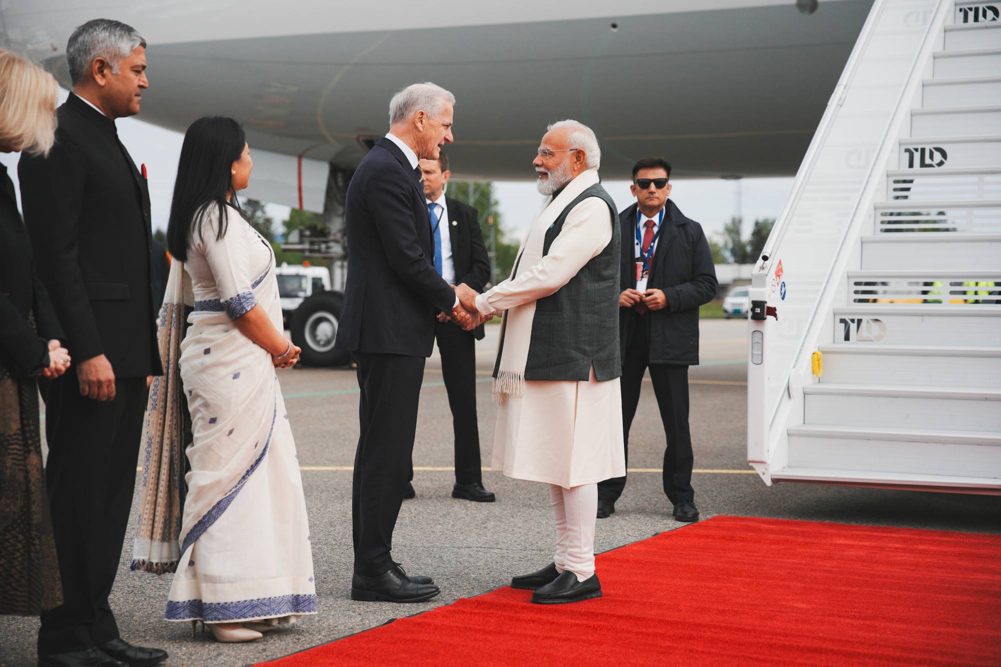Norges statsminister Jonas Gahr Støre ønsker Indias statsminister Narendra Modi velkommen til Norge. De to politikerne håndhilser på en rød løper foran et fly. En kvinne i sari står ved siden av Støre, mens andre personer er bak dem. (Bildebeskrivelsen er laget av en KI-tjeneste)