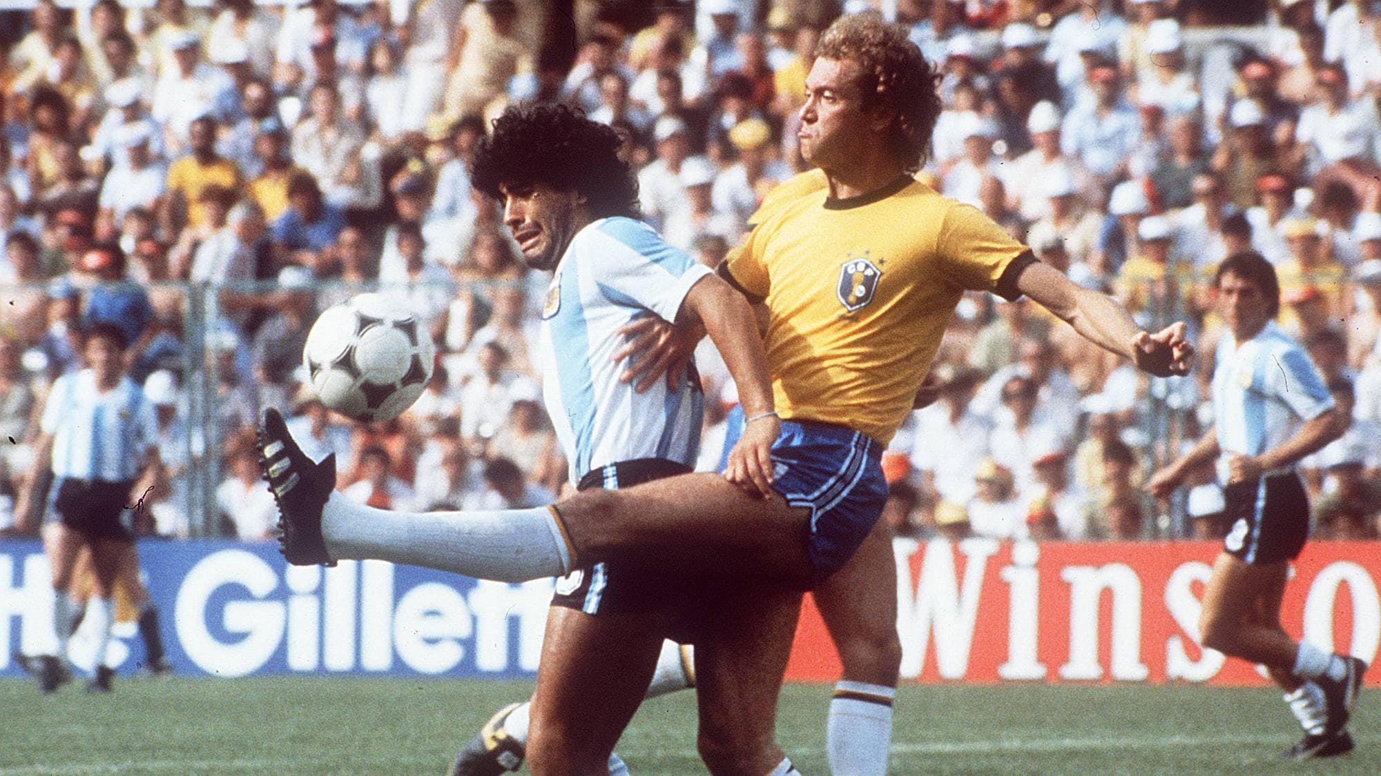 Diego Maradona i duell med en brasiliansk spiller under en fotballkamp. Publikumsfelt i bakgrunnen. (Bildebeskrivelsen er laget av en KI-tjeneste)