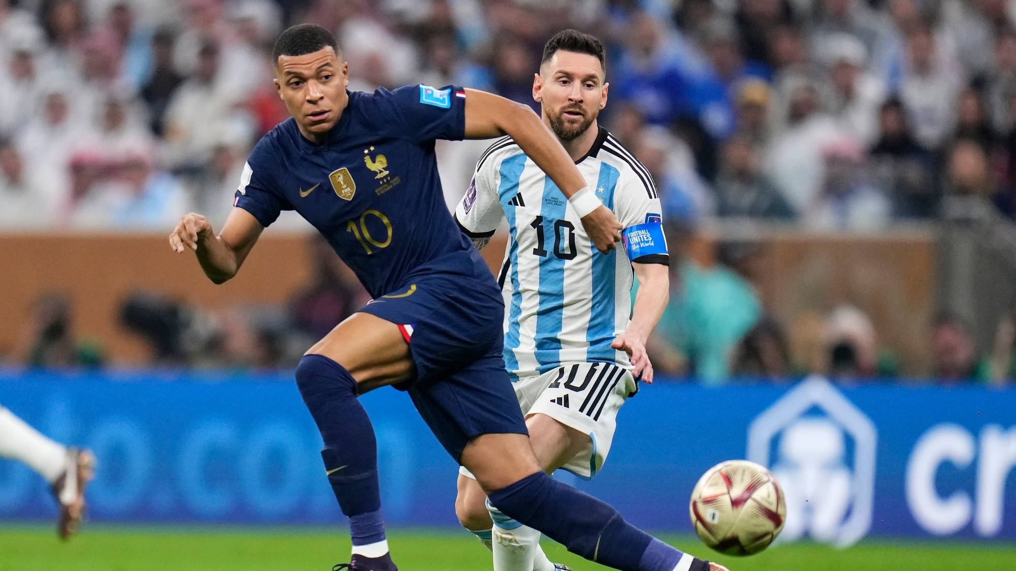 Fotballspillerne Kylian Mbappé (Frankrike) og Lionel Messi (Argentina) er i duell om ballen. De spiller på en grønn gressbane mens publikum sitter bak dem. 