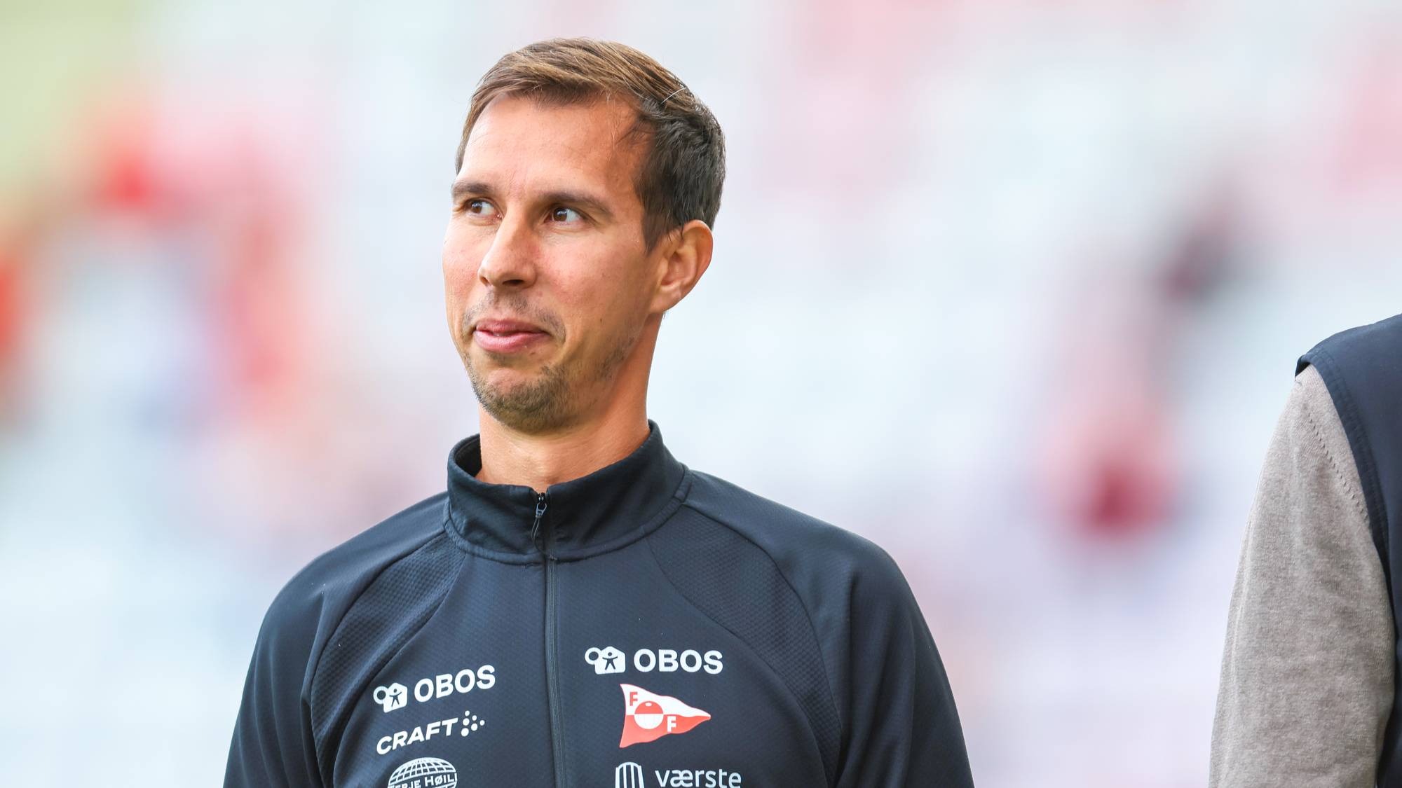 Mannen i mørk treningsgenser med Fredrikstad FK-logo ser til siden. Bakgrunnen er uklar. (Bildebeskrivelsen er laget av en KI-tjeneste)