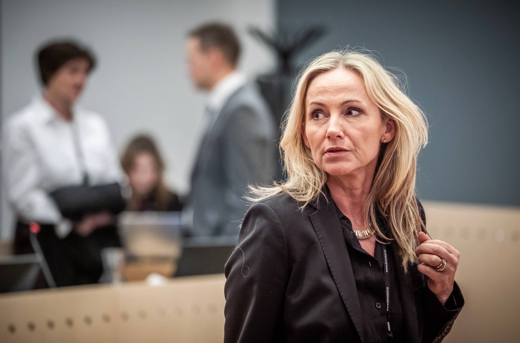 En kvinne med blondt hår i en svart dressjakke ser til siden. Hun har på seg et gullkjede og ringer. I bakgrunnen er det flere utydelige personer i et rettslokale. (Bildebeskrivelsen er laget av en KI-tjeneste)
