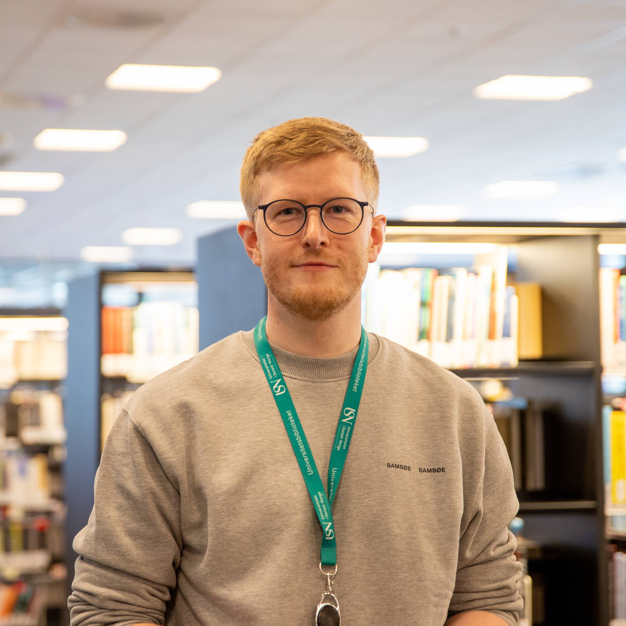En mann på et bibliotek. Han har briller og lys genser.