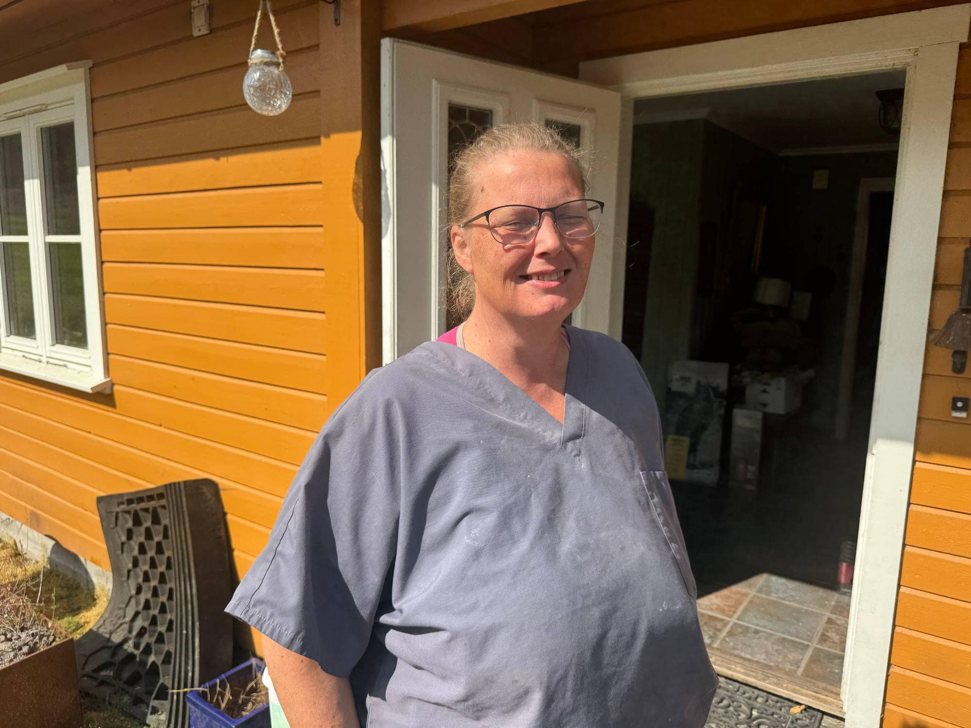 Kvinne med briller og grå overdel står foran en gul trevegg ved en inngangsdør. (Bildebeskrivelsen er laget av en KI-tjeneste)