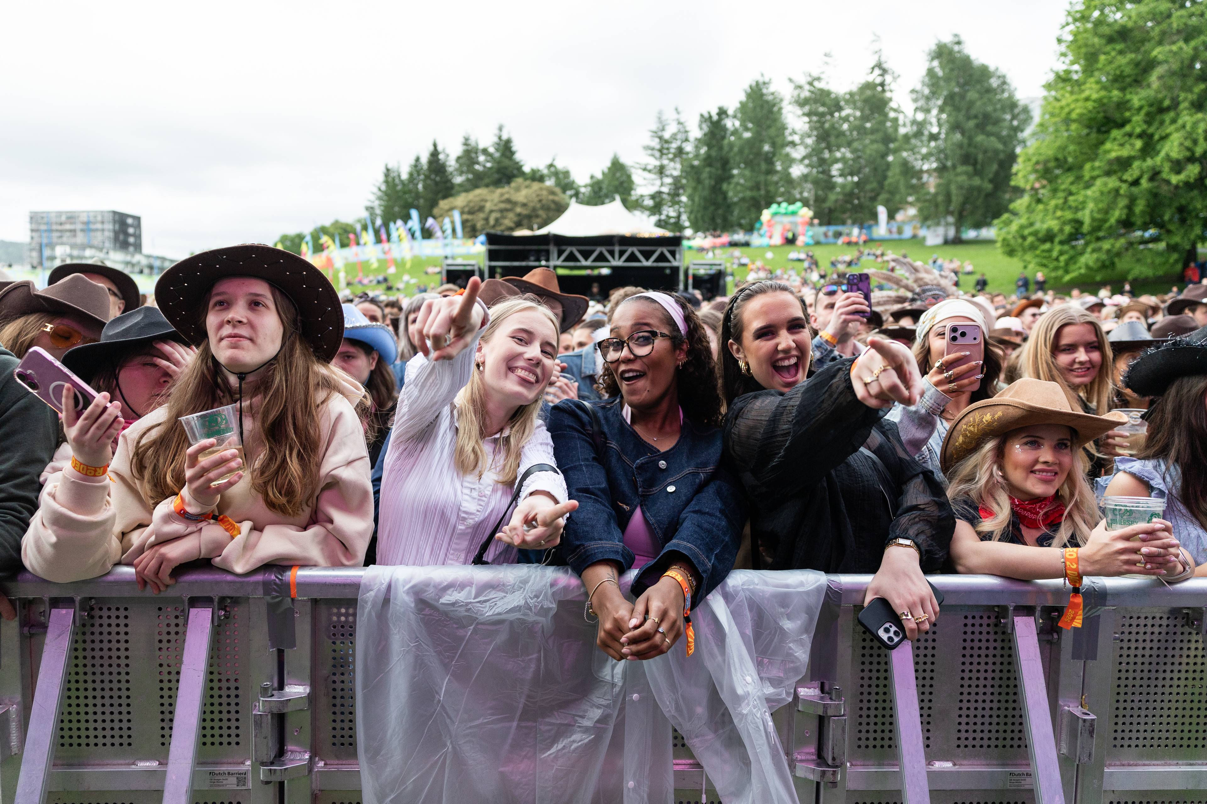 En gruppe unge mennesker med cowboyhatter ser ut til å kose seg på en konsert. De står tett inntil et barrieresystem foran en scene. Flere av dem holder opp mobiltelefoner og peker mot noe utenfor bildeutsnittet. En person til venstre holder en plastkopp med drikke. I bakgrunnen er det et festivalområde med telt, trær og andre tilskuere. (Bildebeskrivelsen er laget av en KI-tjeneste)