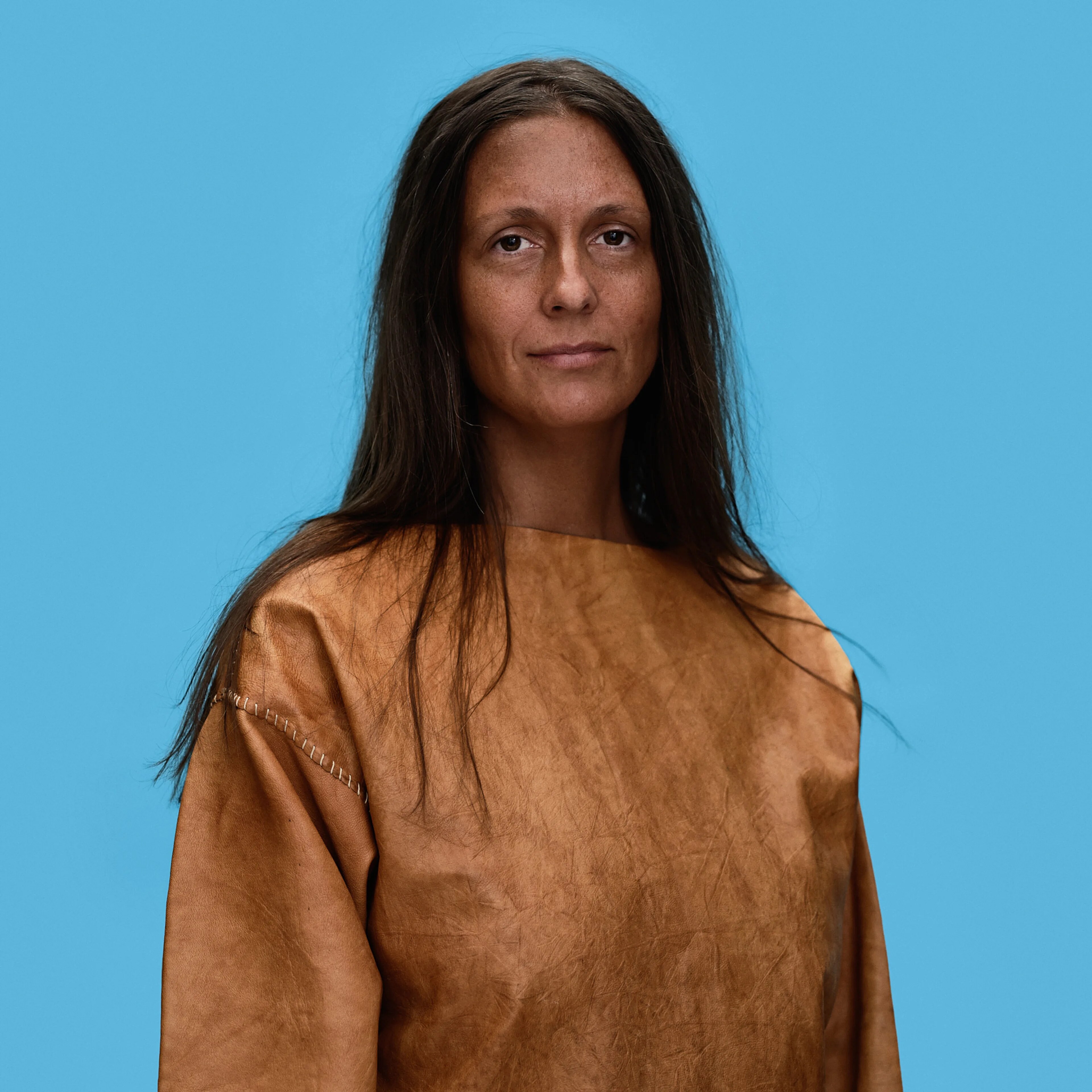 Portrett av en kvinne med langt mørkt hår, iført en brun skinnoverdel mot en lyseblå bakgrunn. (Bildebeskrivelsen er laget av en KI-tjeneste)