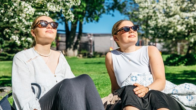 Emma Isdahl og Madelen Lykkebø nyter sola i hagen til Universitetet i Stavanger. Begge sitter med solbriller og myser opp mot sola. I bakgrunnen blomstrer trærne.