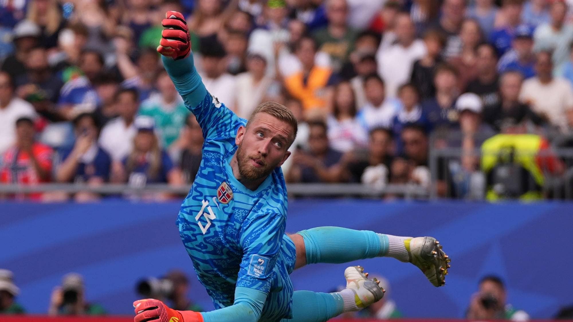 Norsk fotballkeeper i blå drakt kaster seg for å redde ballen foran publikum på tribunen. (Bildebeskrivelsen er laget av en KI-tjeneste)
