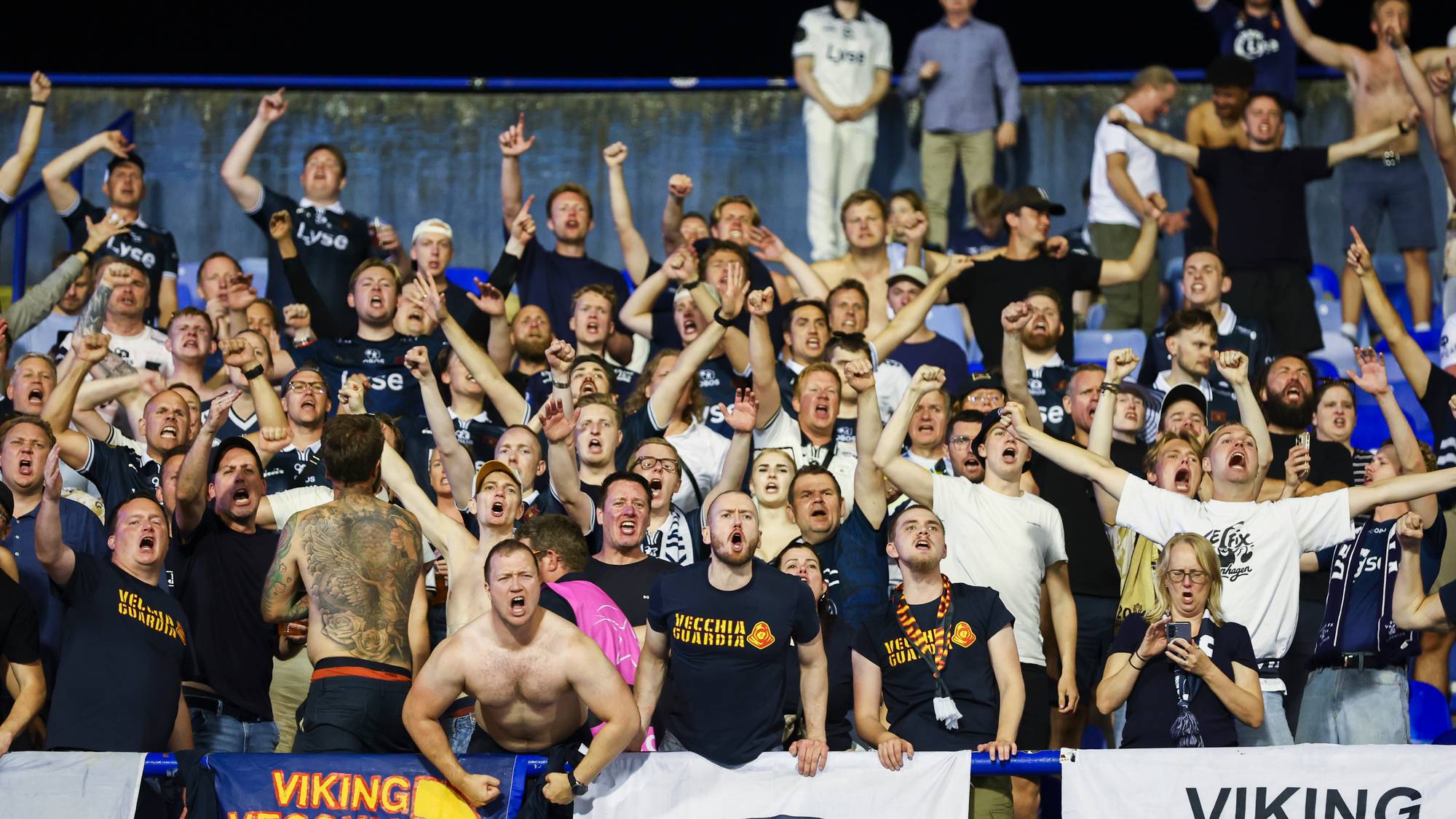 Viking FK-supportere på tribunen, mange med armene i været og ropende. (Bildebeskrivelsen er laget av en KI-tjeneste)