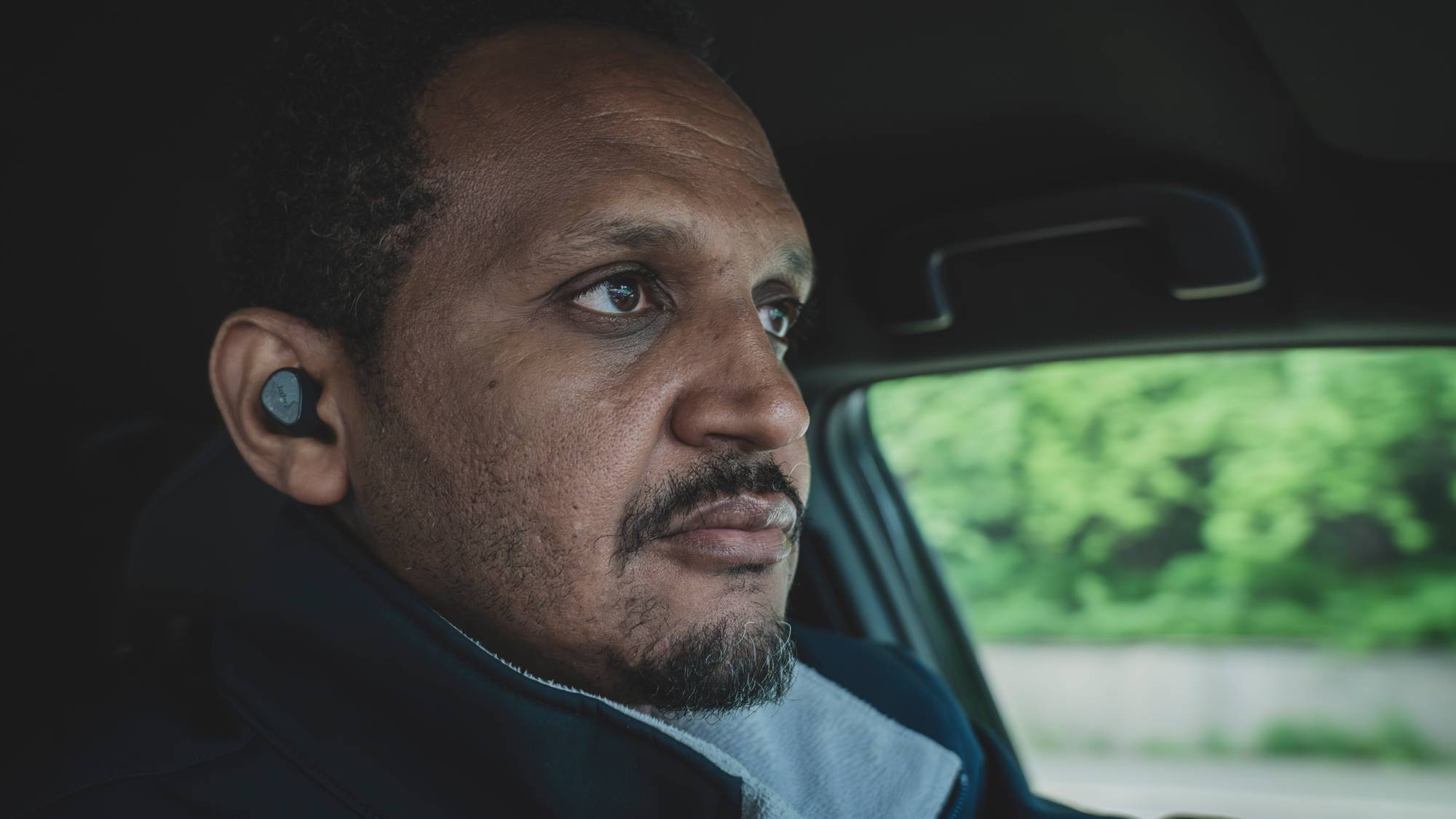 En mann sitter i førersetet på en bil. Han har på seg en mørk jakke og har en øreplugg i venstre øre. Utenfor bilen skimtes grønne trær. (Bildebeskrivelsen er laget av en KI-tjeneste)