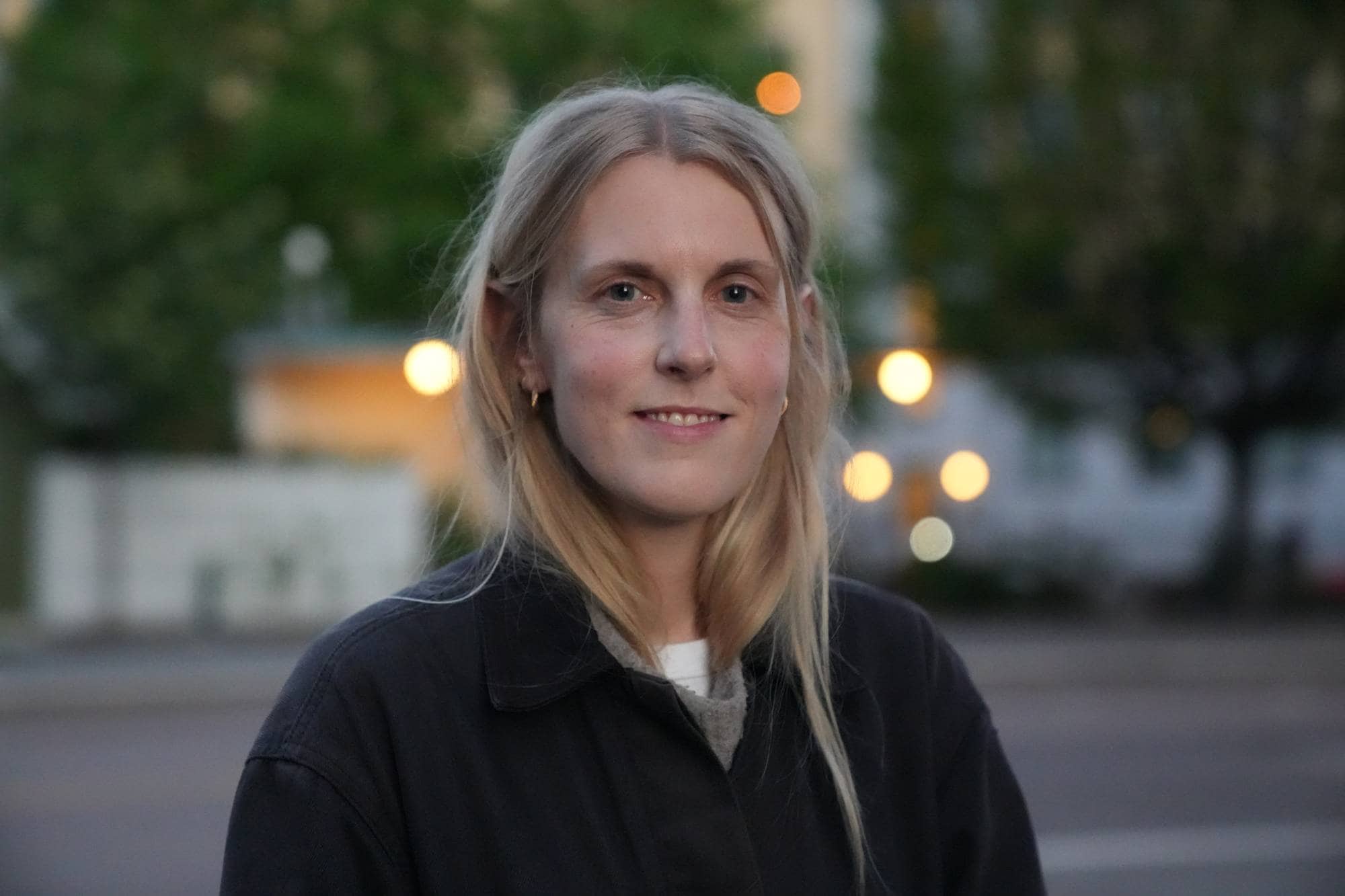 En ung kvinne med blondt hår og en mørk jakke smiler mot kameraet. Bak henne er det utydelige bygninger og trær. Lyset fra lyktestolper skaper uklare, gule sirkler i bakgrunnen. (Bildebeskrivelsen er laget av en KI-tjeneste)