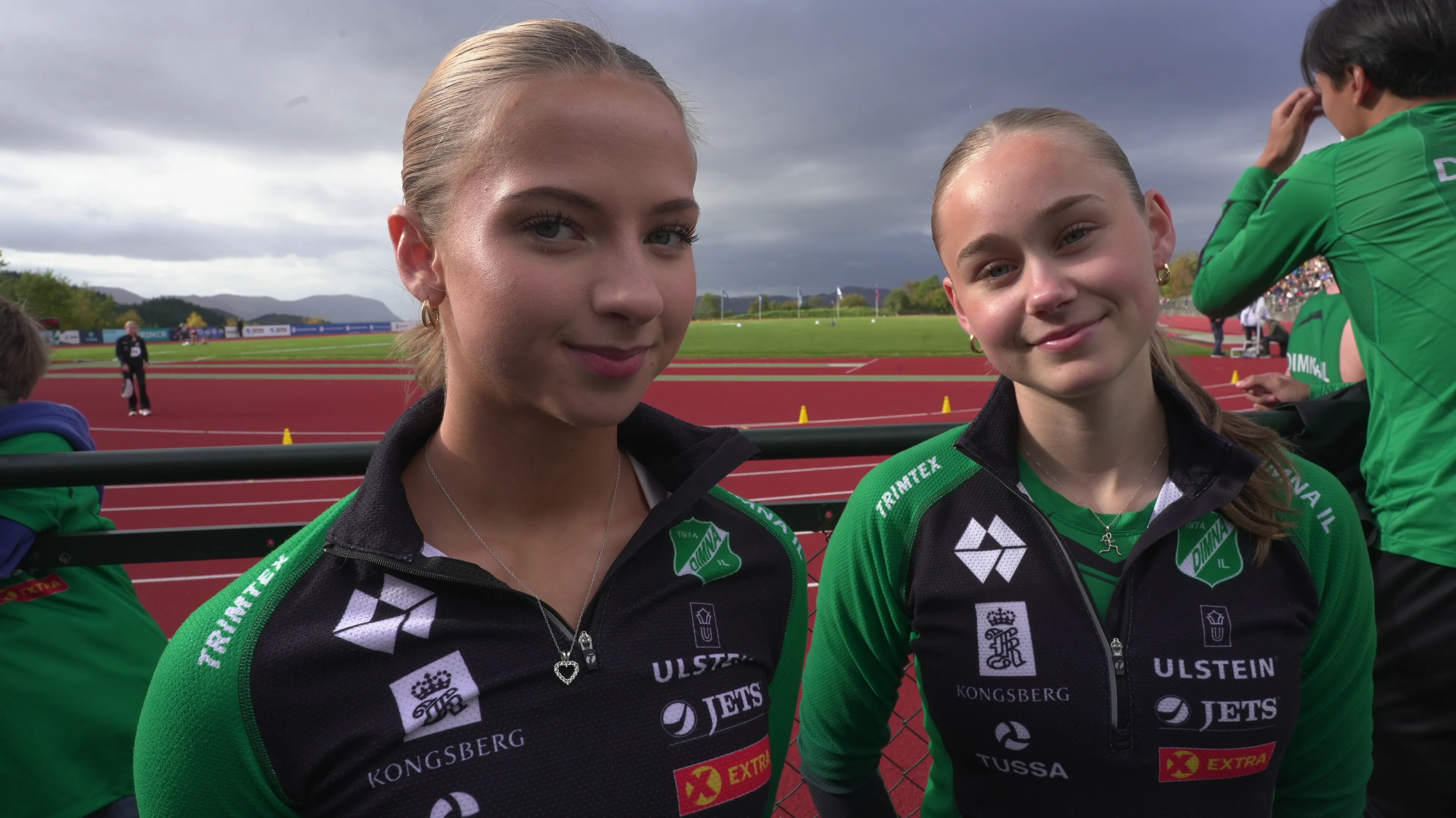 To unge kvinner i klubbdrakt står på en friidrettsbane med løpebaner i bakgrunnen. (Bildebeskrivelsen er laget av en KI-tjeneste)