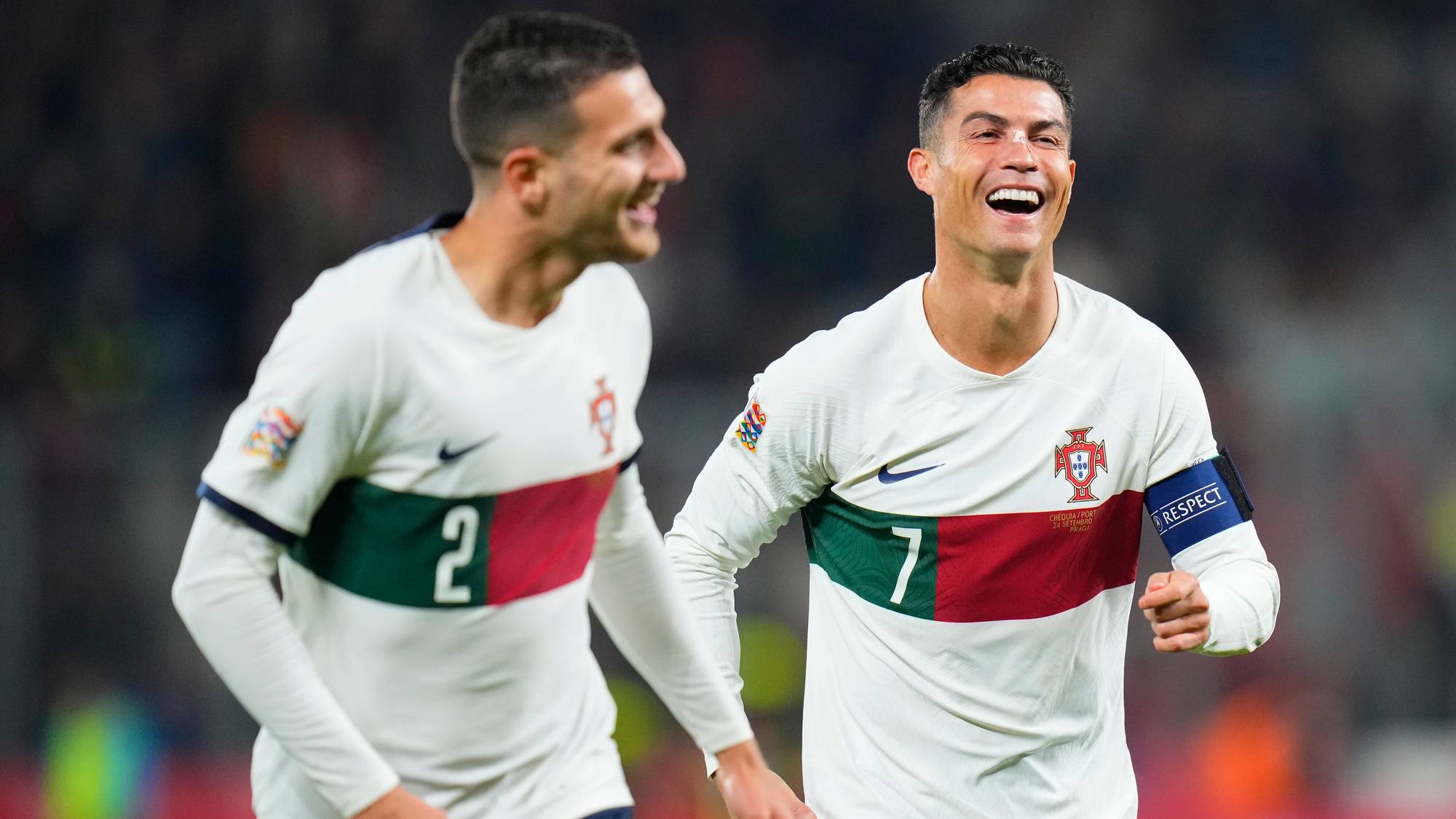 Two men in white football jerseys with red and green stripes are smiling. One has the number 2 on his jersey, and the other has the number 7. They appear to be on a football field. (Bildebeskrivelsen er laget av en KI-tjeneste)