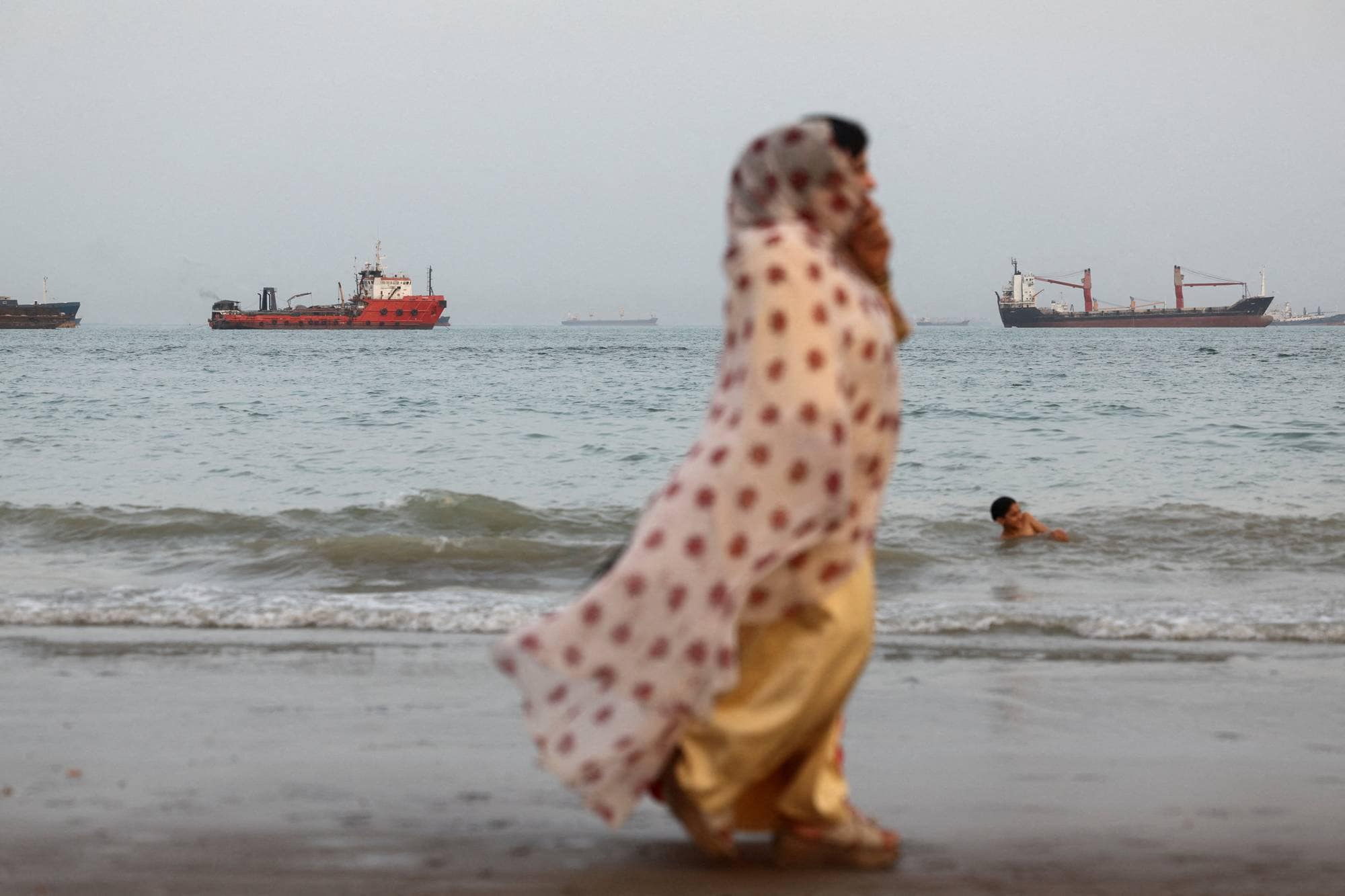 Lasteskip i Hormuzsundet. I forgrunnen ei kvinne i sjal som går på en strand.
