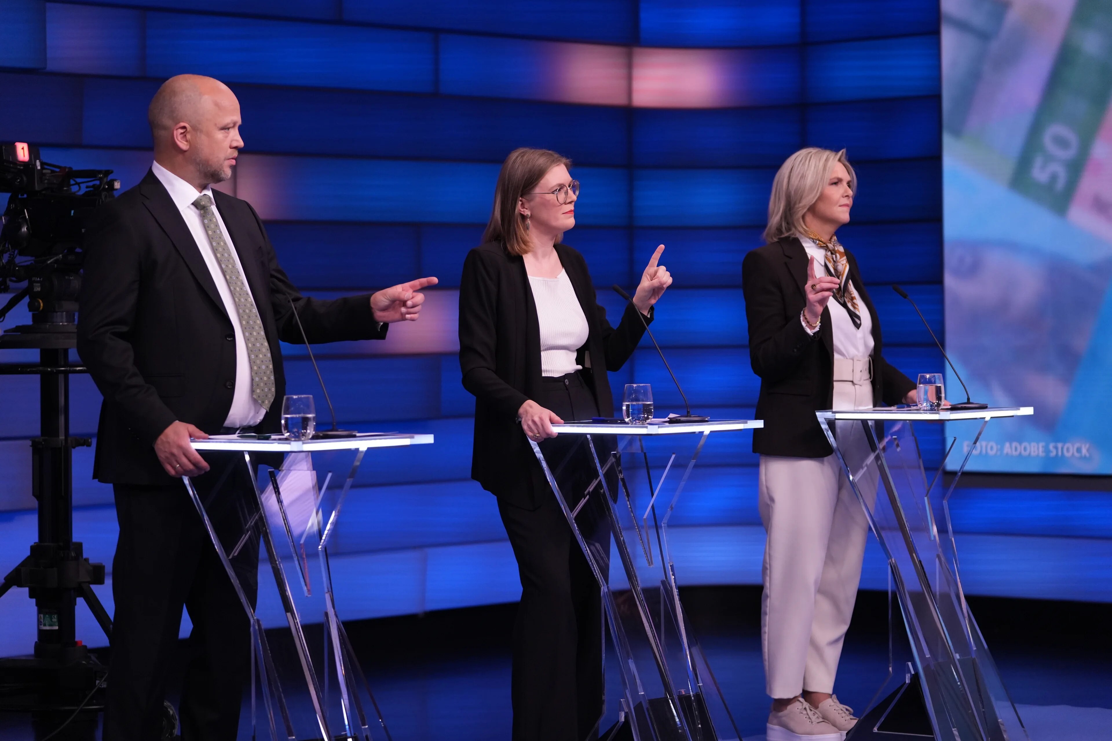 Tre personer står ved hver sin talerstol i et TV-studio foran en blå skjerm. (Bildebeskrivelsen er laget av en KI-tjeneste)