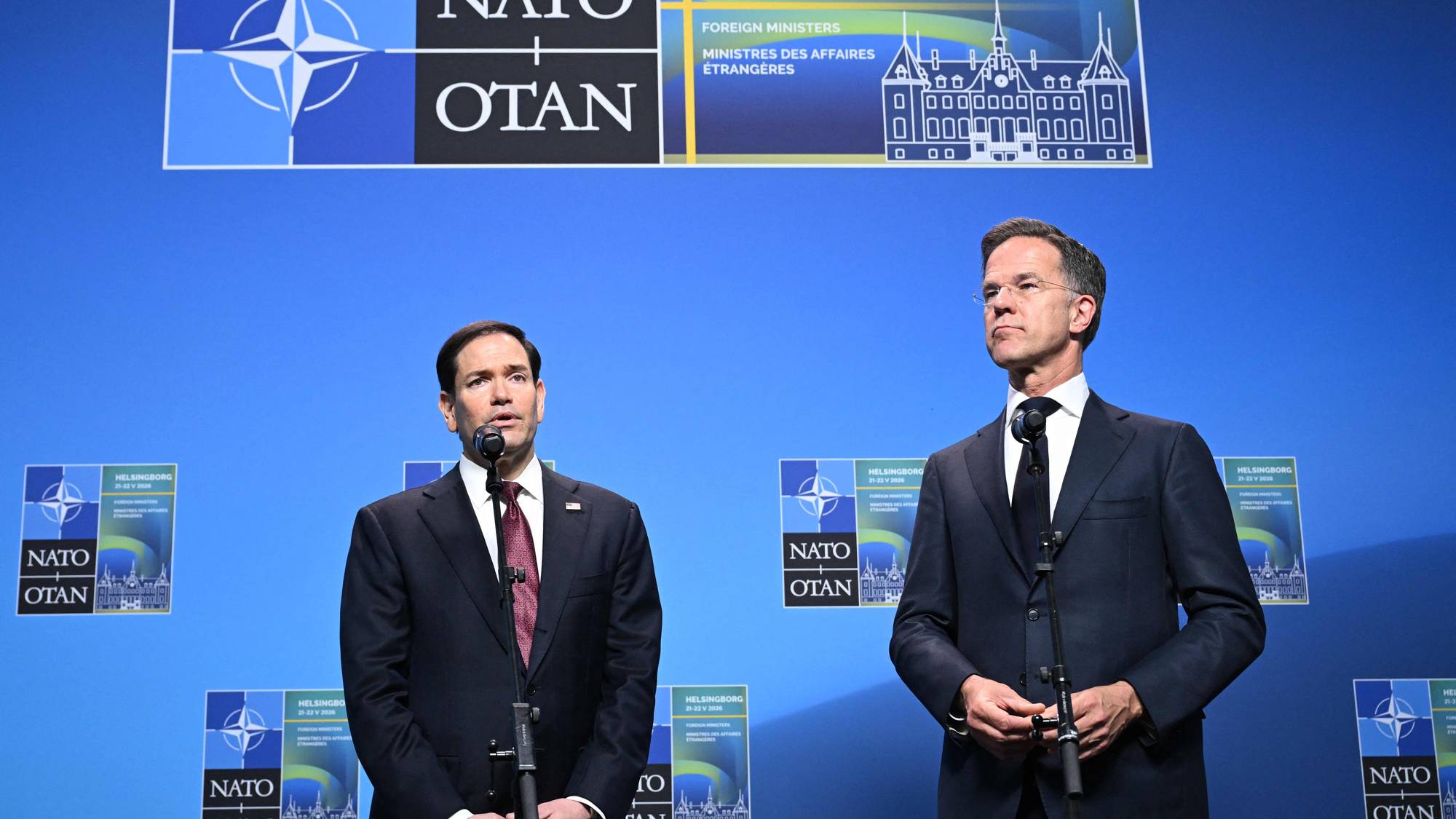 To menn i dress står foran en blå bakgrunn med NATO-logo. Begge er plassert bak talerstoler med mikrofoner.Bak dem er det store bannere med NATO-logo og informasjon om Helsingborg Foreign Ministers. (Bildebeskrivelsen er laget av en KI-tjeneste)