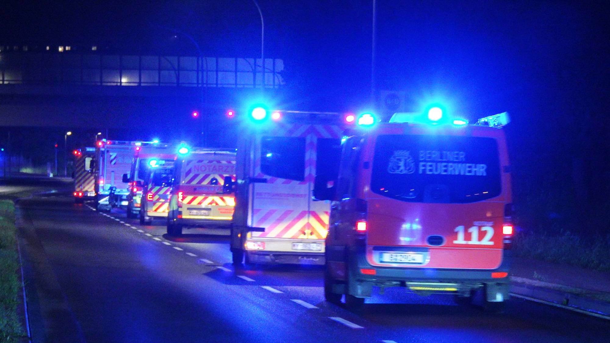 Ambulanser og brannbiler med blålys står på en vei om natten. En av bilene er merket med "Berliner Feuerwehr". Det er en overliggende struktur synlig i bakgrunnen, og lys fra trafikklys og biler. (Bildebeskrivelsen er laget av en KI-tjeneste)