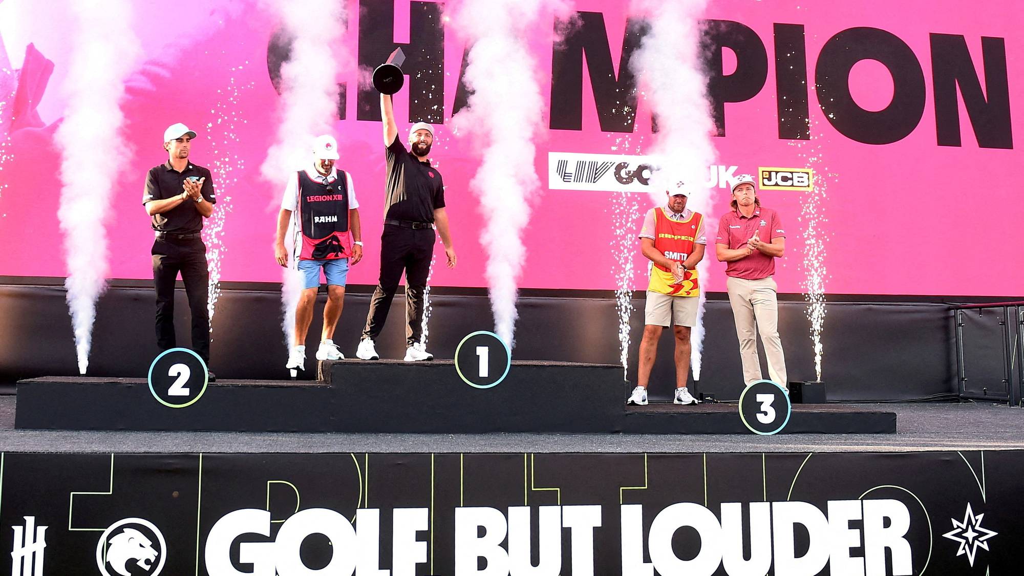 Fem personer står på en scene med røyk og konfetti. Midt på scenen står en mann med trofé over hodet. Bak dem er det en rosa bakgrunn med tekst. Langs scenekanten er det et skilt med teksten "GOLF, BUT LOUDER". (Bildebeskrivelsen er laget av en KI-tjeneste)