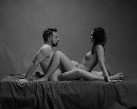 En naken mann og en naken dame sitter på senga med bena rundt hverandre mens de ser hverandre inn i øynene