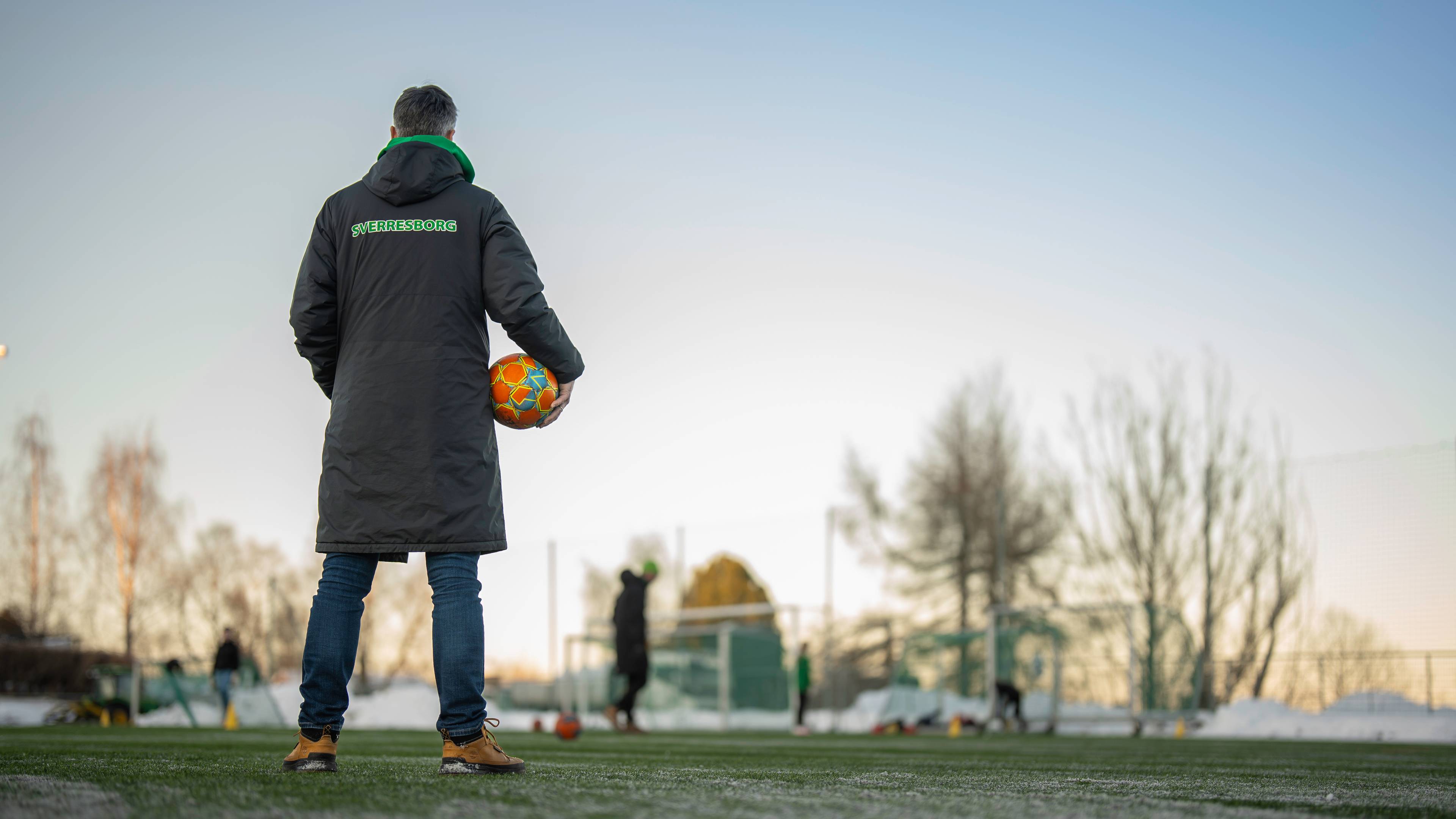 Mann står med ryggen til på en fotballbane, og holder en ball i arma