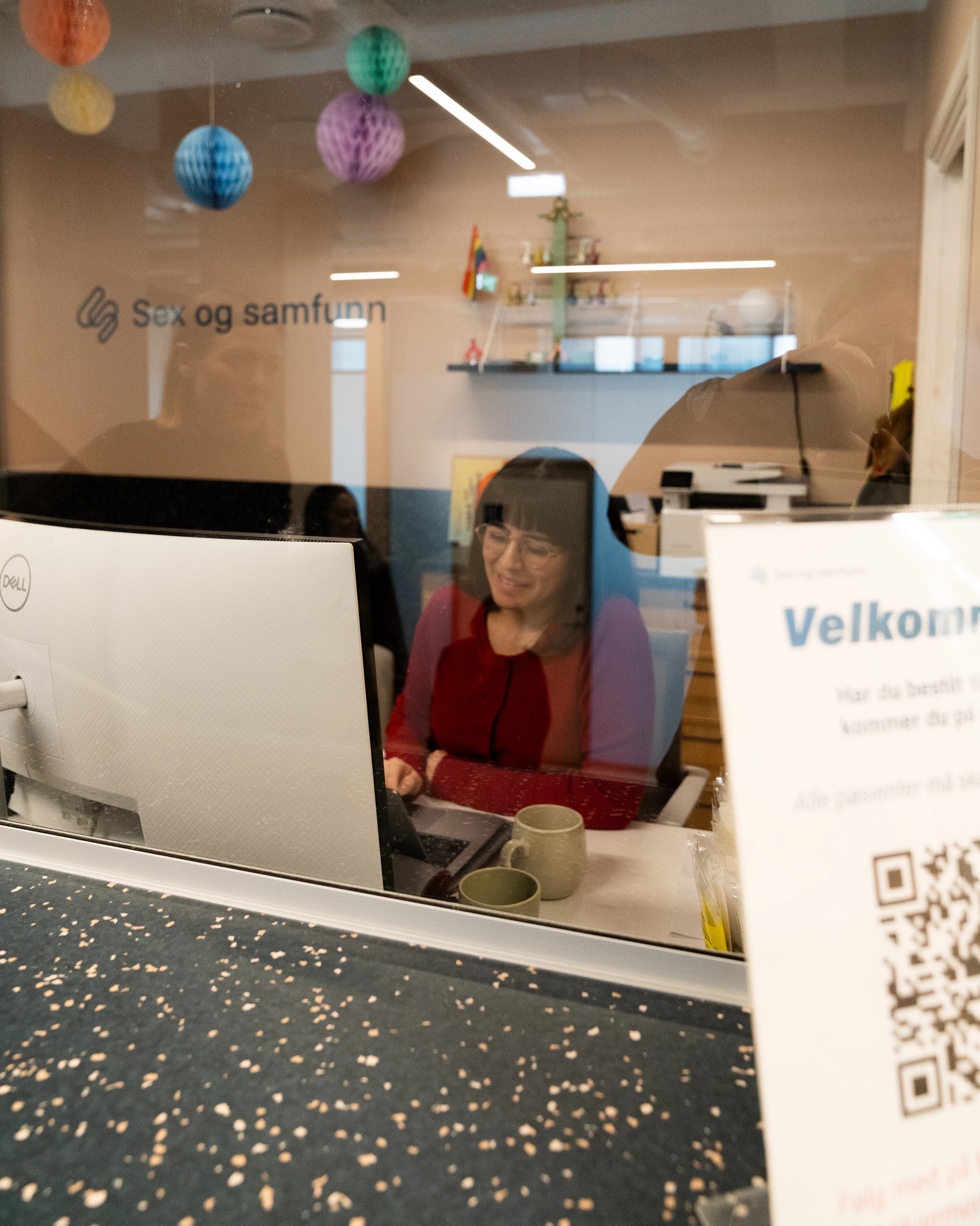 en kvinne sitter i resepsjonen  og smiler, bak står det skrevet sex og samfunn på veggen