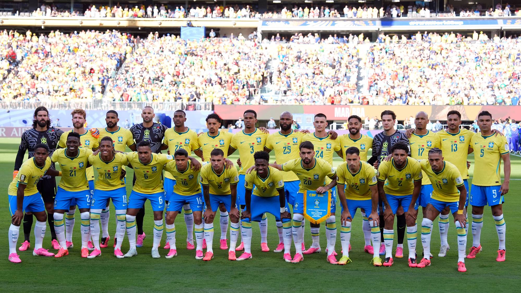 Brasil-landslaget i fotball poserer for bilde før kamp. De står oppstilt på en gressbane med tribuner fulle av tilskuere i bakgrunnen. Spillerne er kledd i gule trøyer og blå shorts. (Bildebeskrivelsen er laget av en KI-tjeneste)