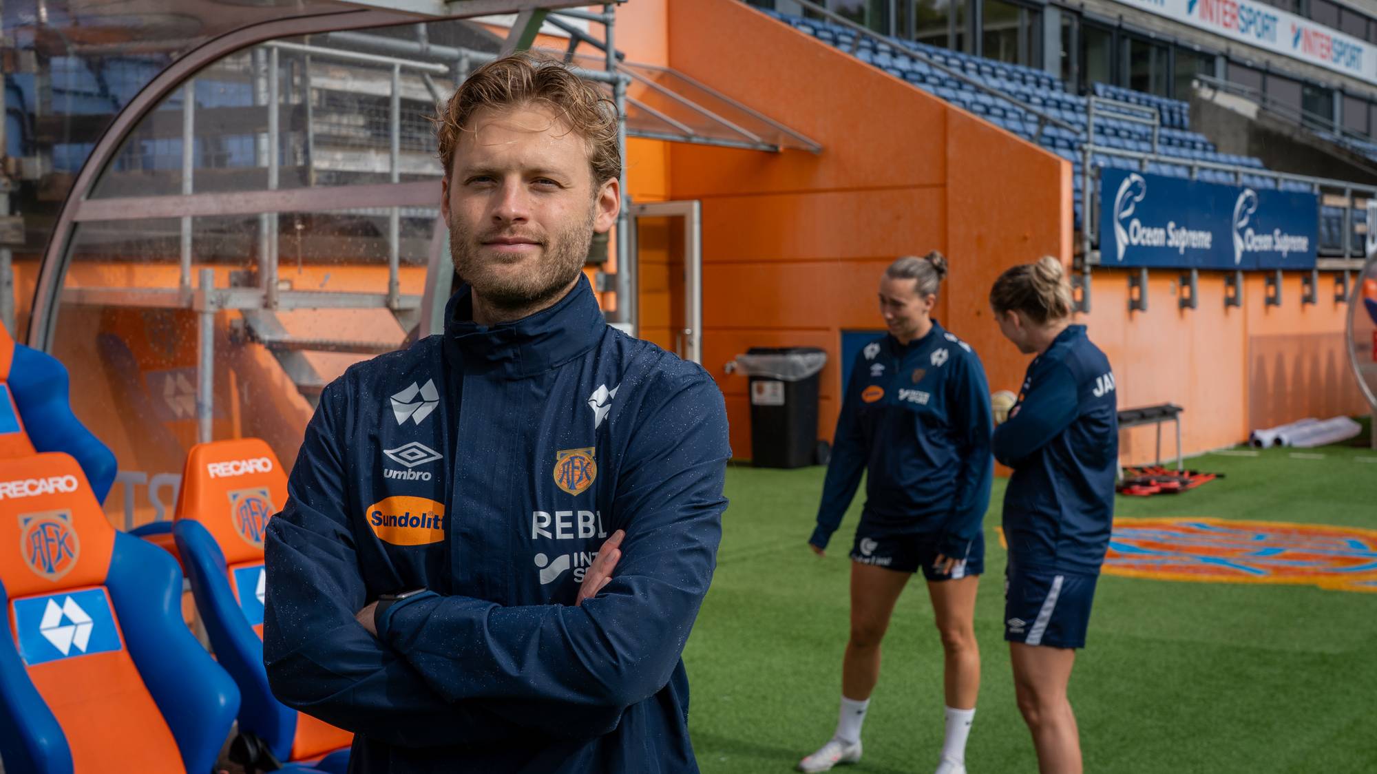 En mann i Aalesund-jakke står på sidelinjen med to spillere i bakgrunnen på en fotballstadion. (Bildebeskrivelsen er laget av en KI-tjeneste)