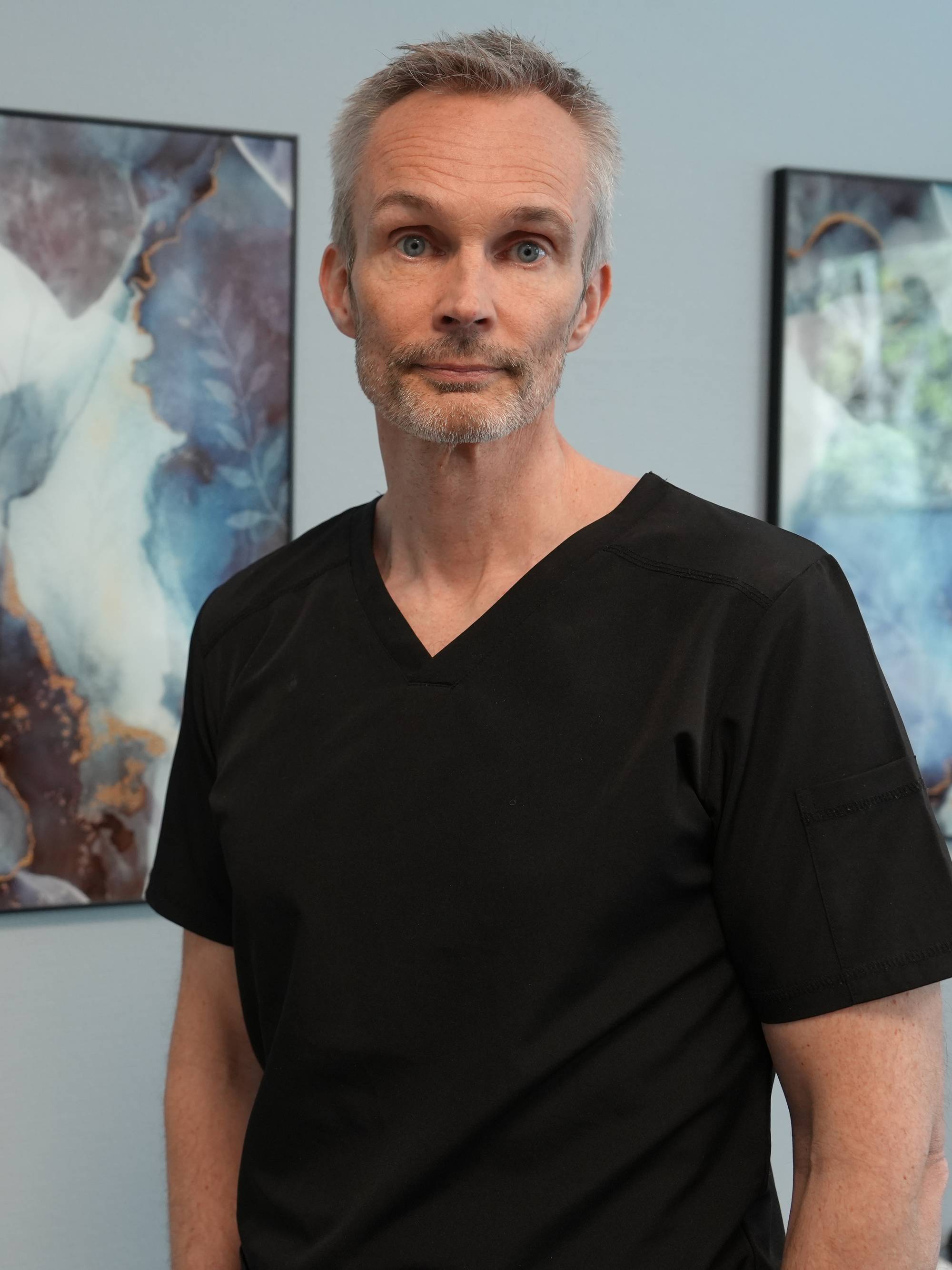 Ein mann i svart t-skjorte står framfor to fargerike kunstverk og ser alvorleg ut. 