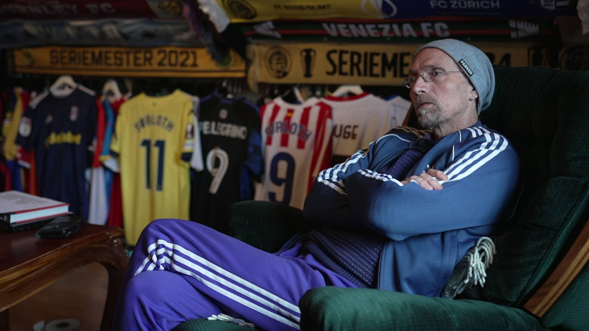 En mann sitter i en lenestol med armene i kors. Bak ham henger flere fotballdrakter og supporter-skjerf. Mannen har grå lue, briller og et blått treningsantrekk med hvite striper. (Bildebeskrivelsen er laget av en KI-tjeneste)