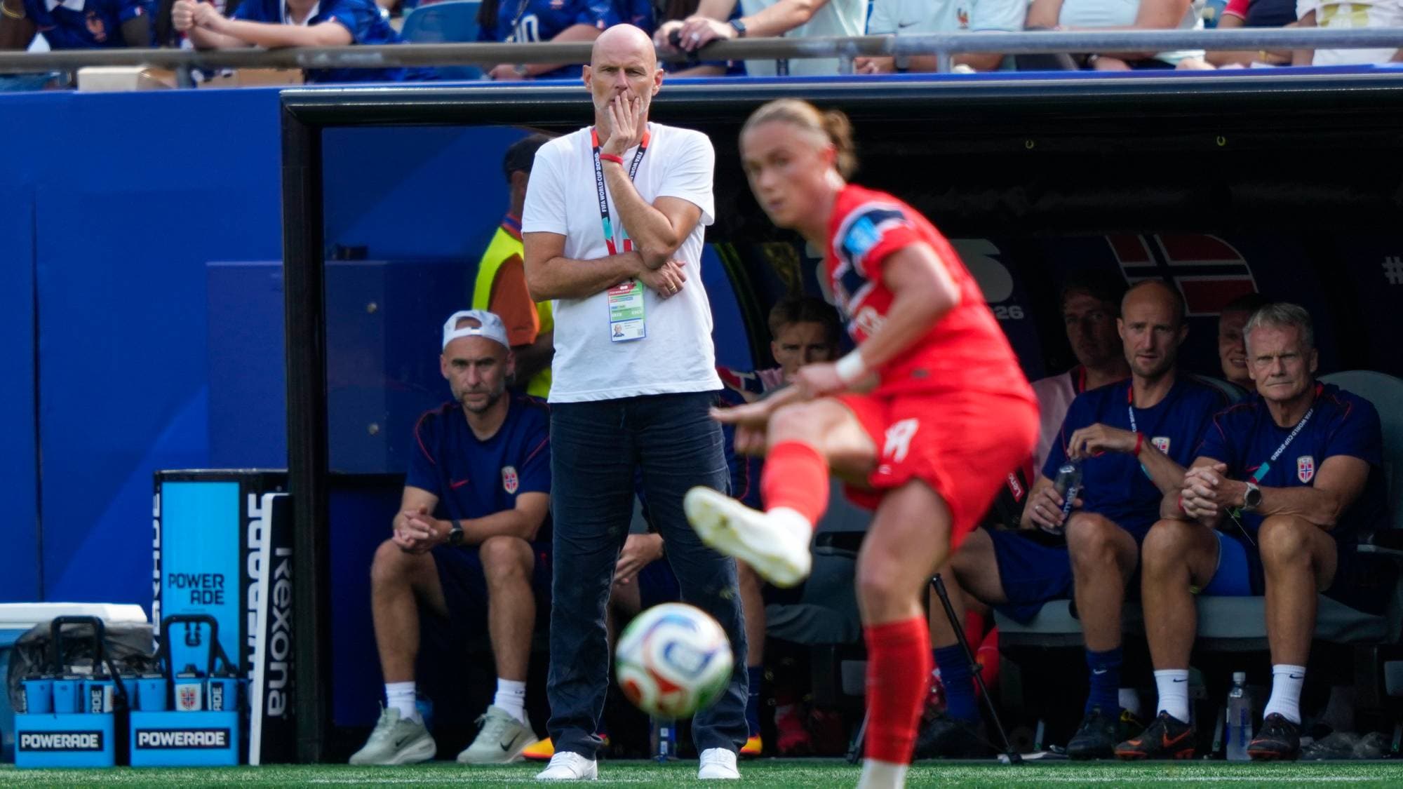 Trener Hege Riise ser på en spiller som skyter ballen foran innbytterbenken til Norge. (Bildebeskrivelsen er laget av en KI-tjeneste)