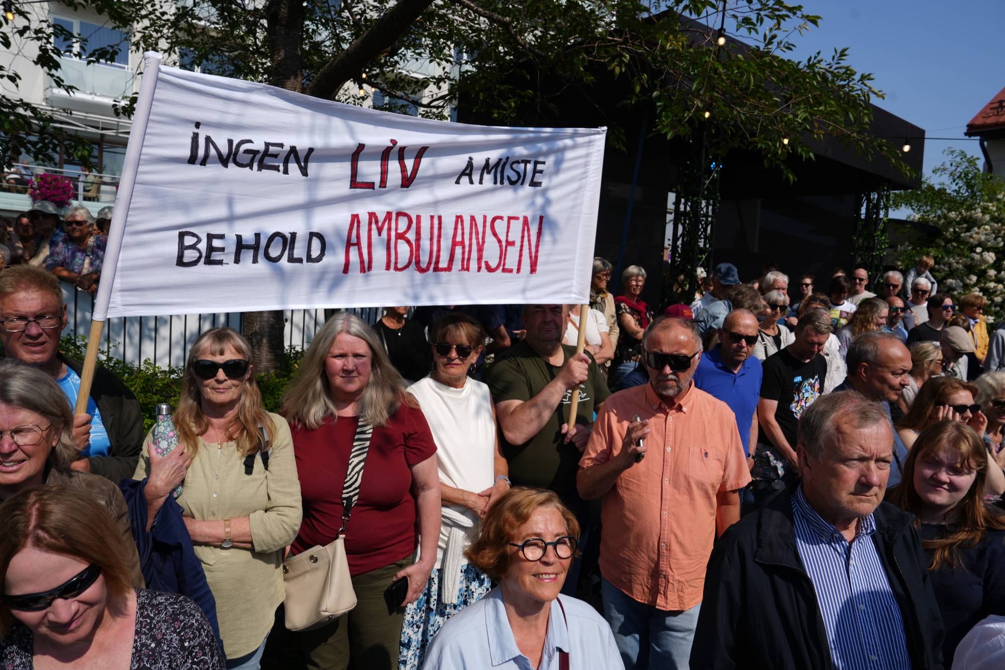 Folk demonstrerer med et banner hvor det står Ingen liv å miste, behold ambulansen. (Bildebeskrivelsen er laget av en KI-tjeneste)
