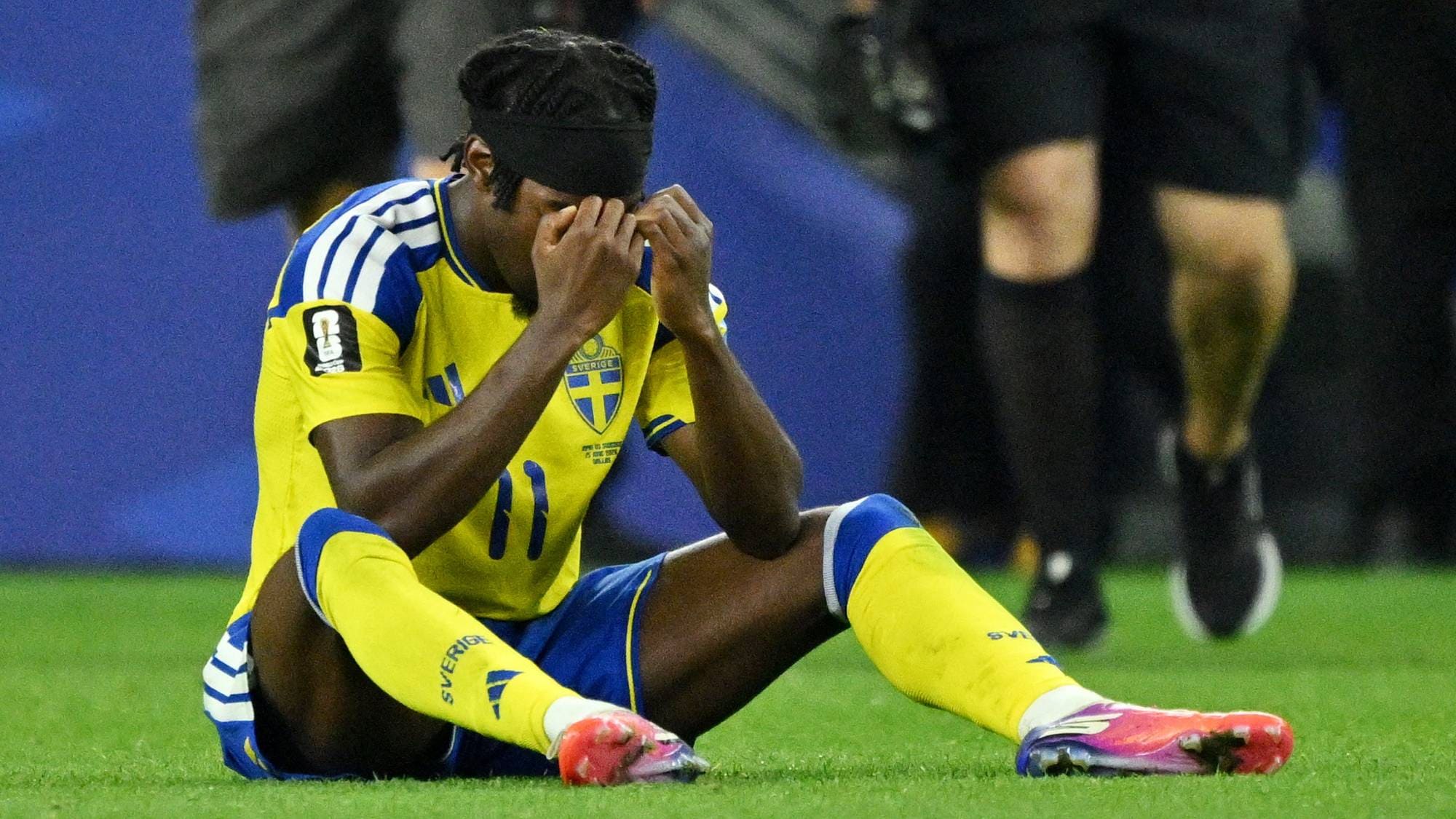 Svensk fotballspiller sitter på gresset og holder seg til ansiktet etter en kamp. (Bildebeskrivelsen er laget av en KI-tjeneste)