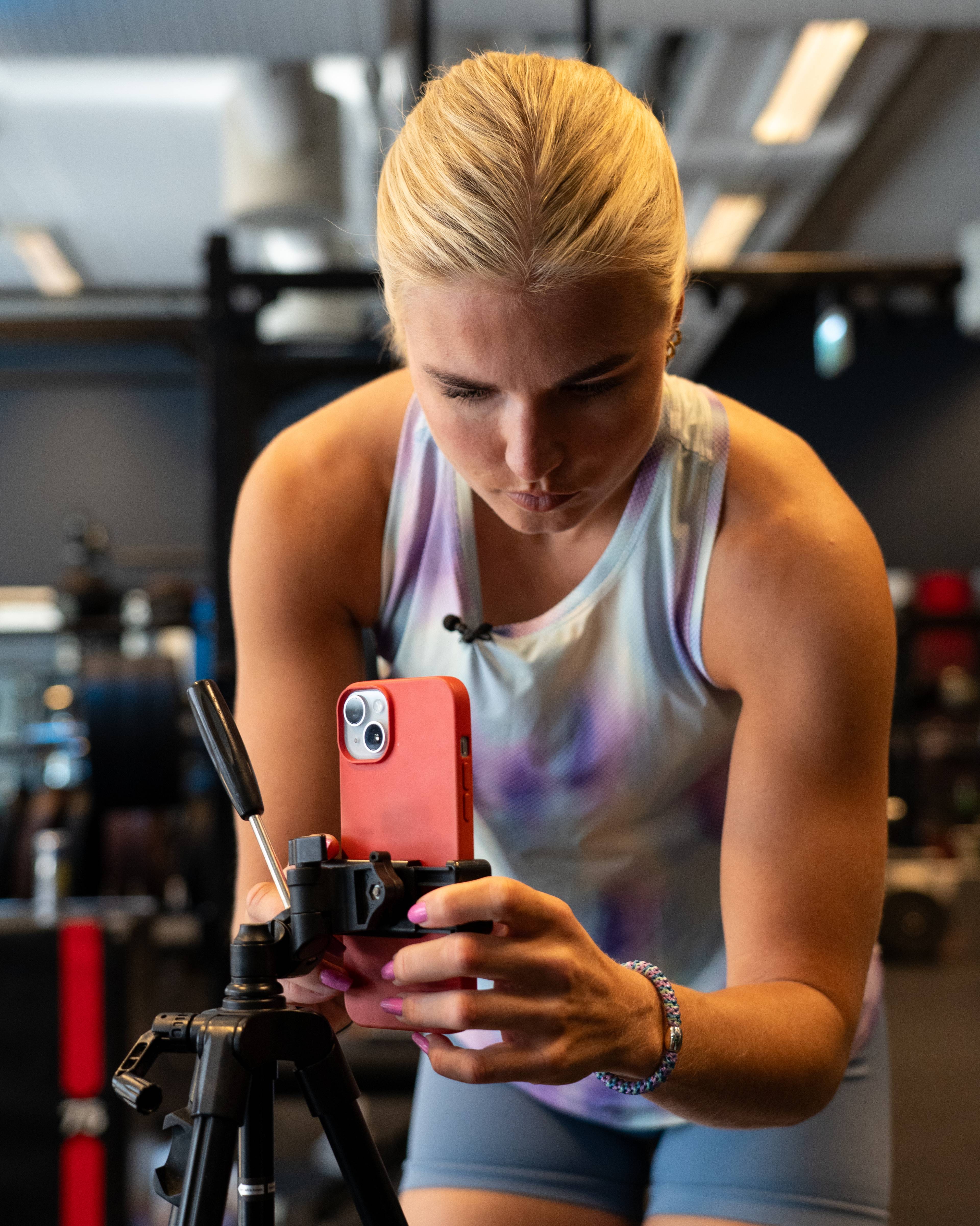 En kvinne med blondt hår filmer seg selv med en mobiltelefon. Hun står i et treningsstudio med en tripod foran seg. Kvinnen er iført en trenings-topp og shorts. (Bildebeskrivelsen er laget av en KI-tjeneste)