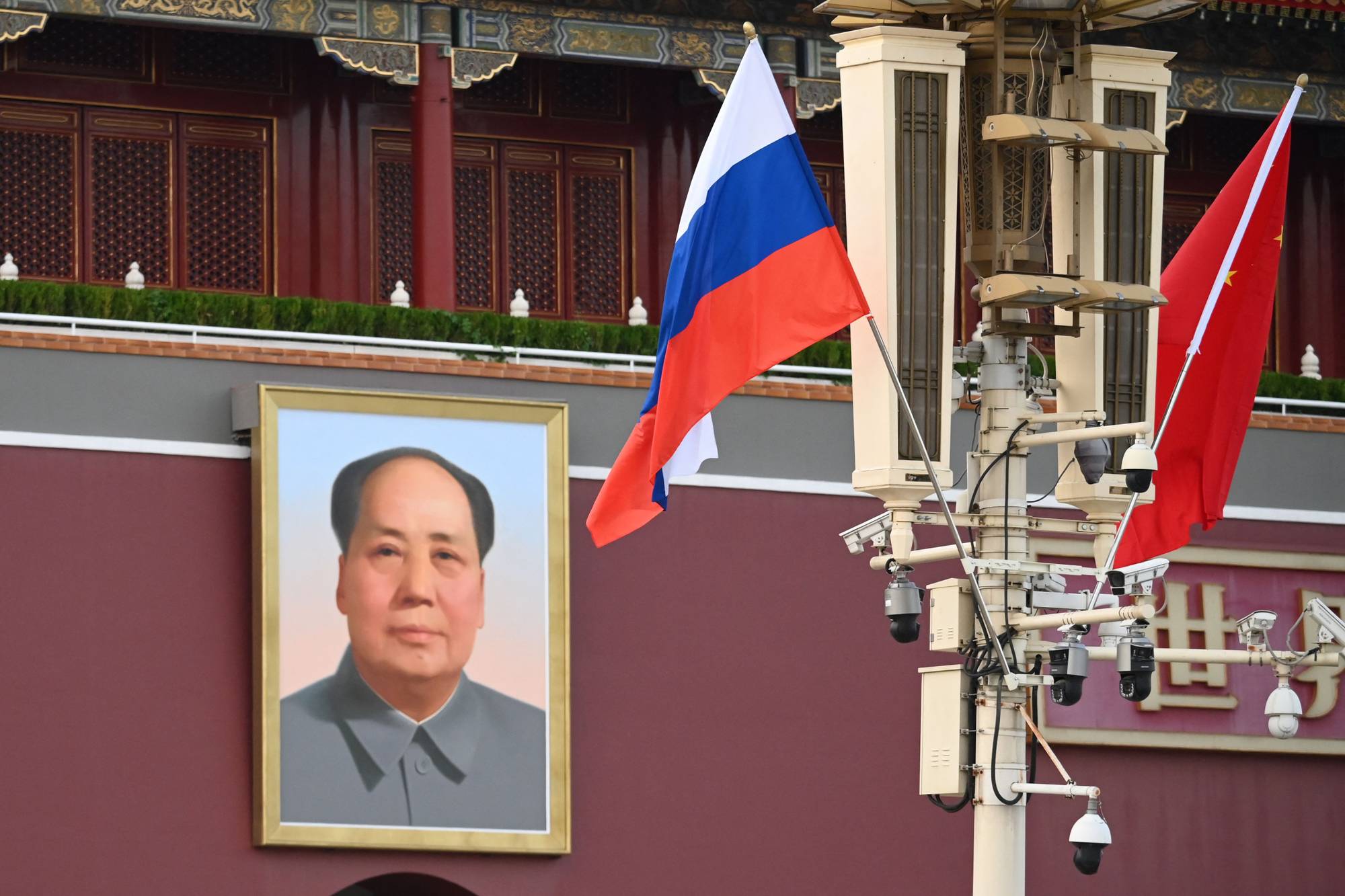 Et portrett av Mao Zedong henger ved siden av det russiske og det kinesiske flagget. En lysmast med flere overvåkningskameraer står foran den rødmalte bygningen. Bygningen har tradisjonelle kinesiske vinduer og et gullfarget tak. (Bildebeskrivelsen er laget av en KI-tjeneste)