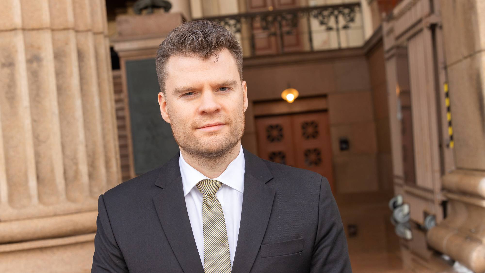 Andreas C. Halse, Advokat i Elden advokatfirma, foran en bygning med murstein og store stolper. Advokaten har på seg en svart dress med hvit skjorte og slips. Halse ser rett mot kamera.